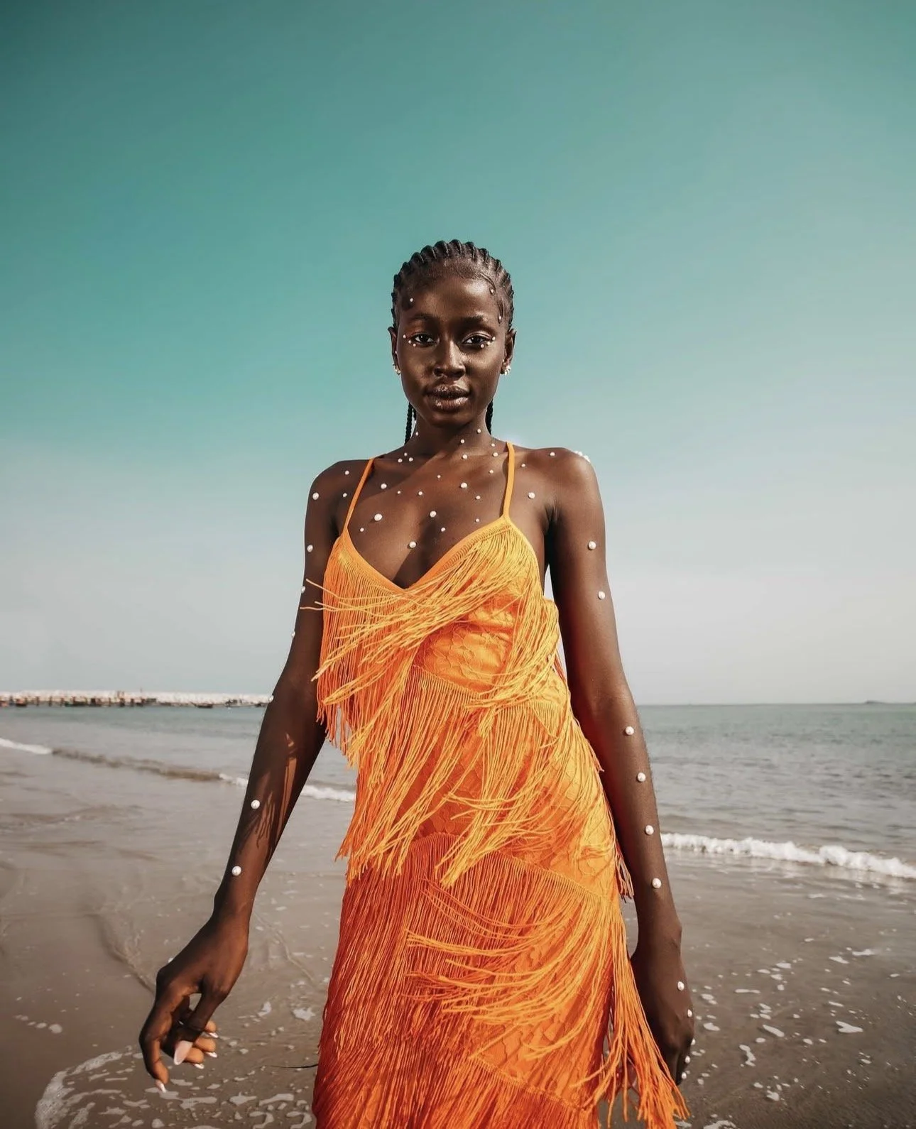 A woman in an orange fringed dress standing on a beach with the ocean in the background.