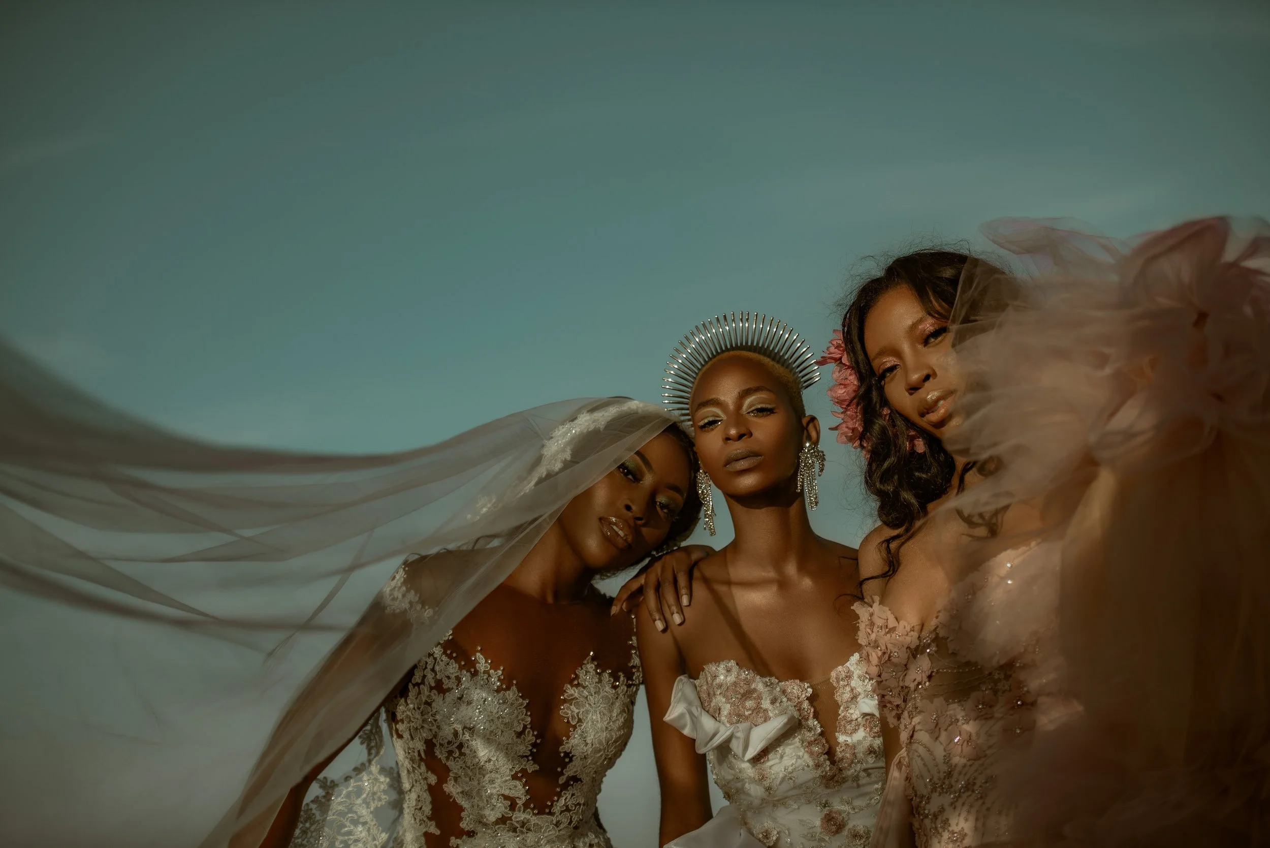 Three women in elegant dresses pose closely together outdoors, with one wearing a crown and veil, and the others with floral accessories, against a clear sky.