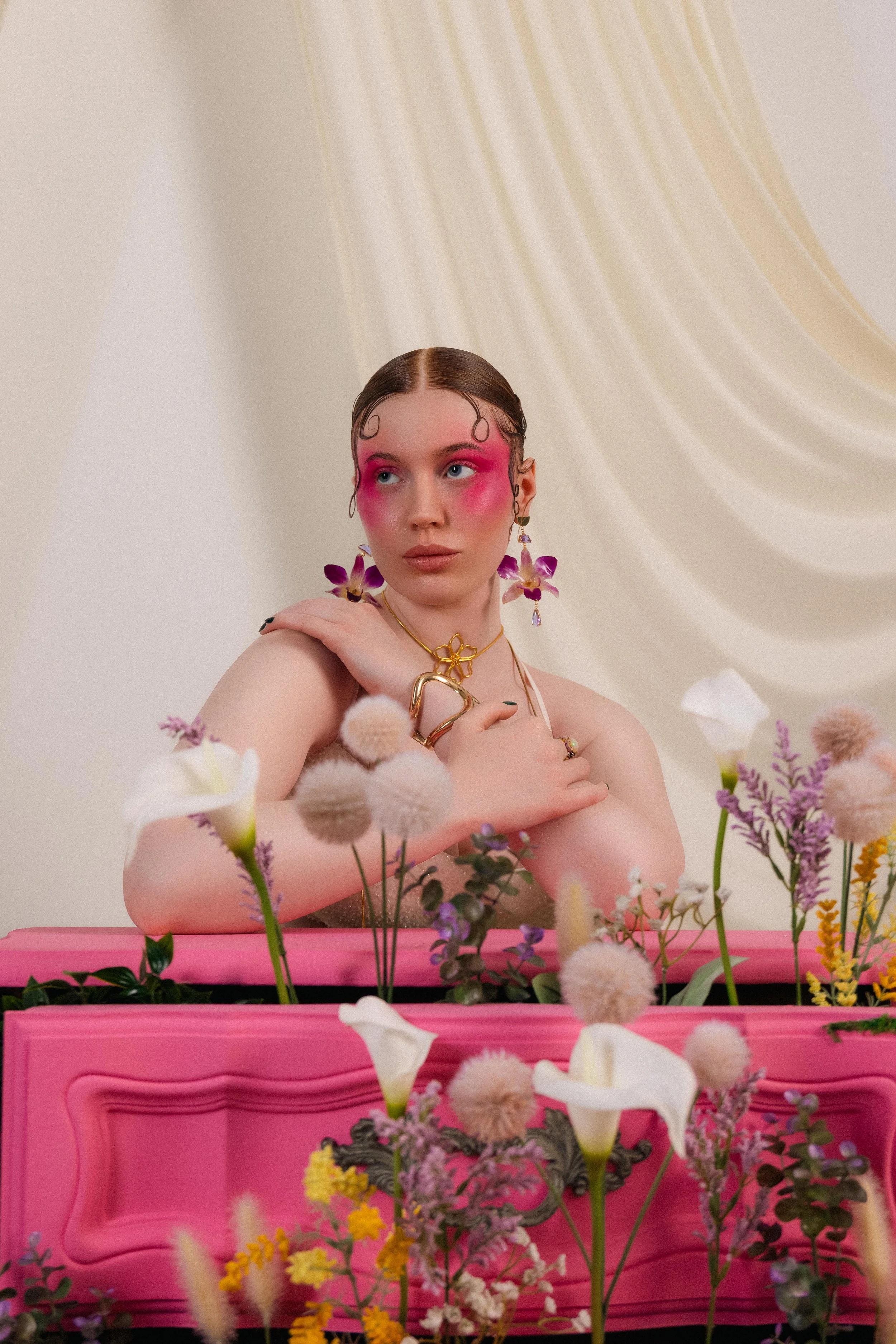 A woman with pink eye makeup and flowers in her hair, wearing jewelry, sitting behind a pink surface adorned with various flowers, against a cream-colored backdrop with draped fabric.