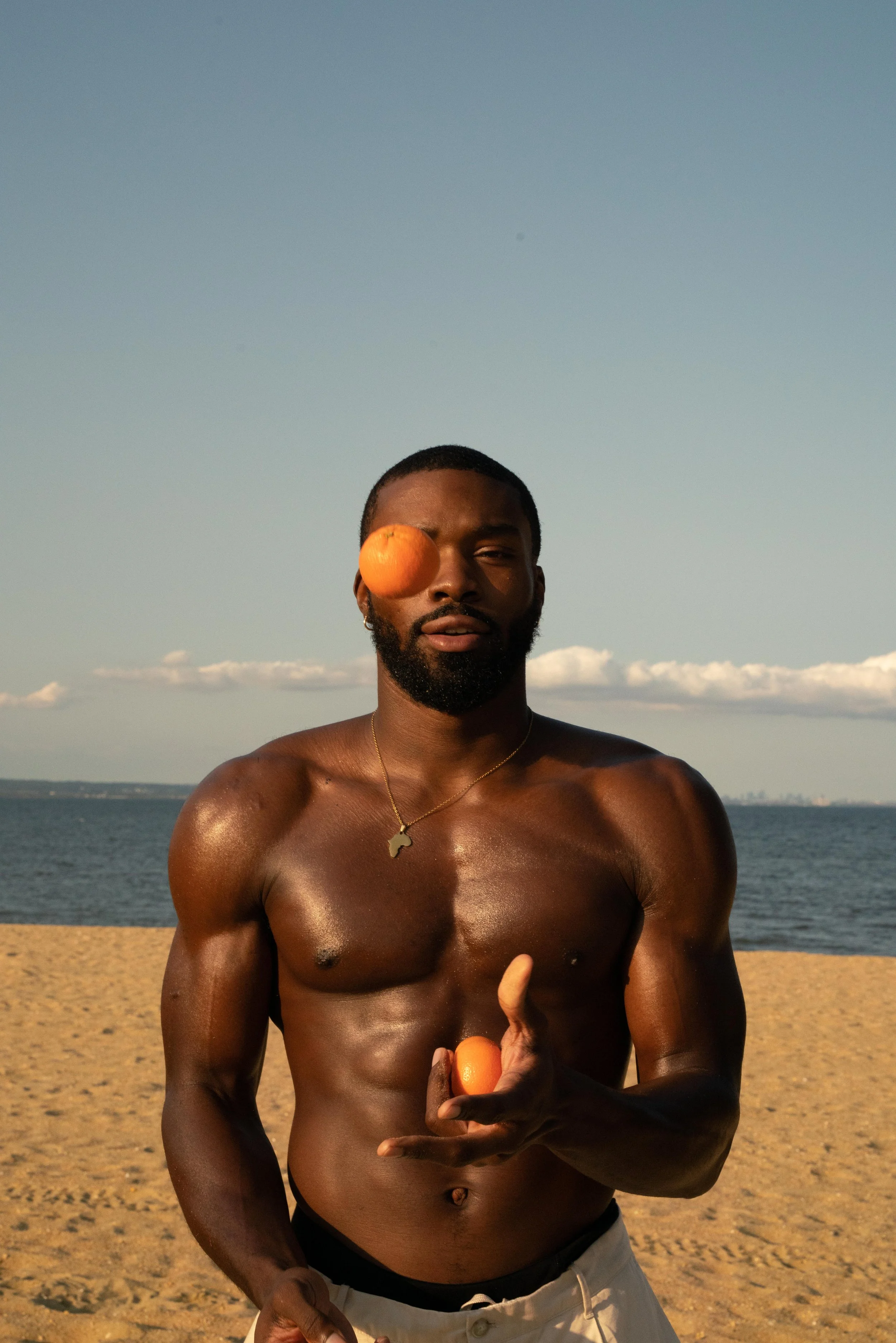 A shirtless man standing on a sandy beach holding a small orange in his hand, with an orange over his left eye, and wearing a gold necklace with an Africa-shaped pendant. The ocean and blue sky with some clouds are in the background.