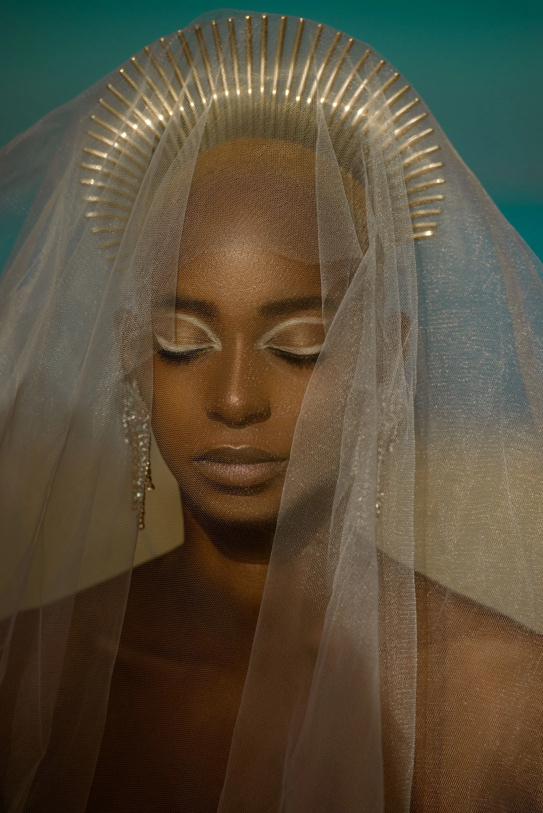 A woman with dark skin and closed eyes, partially covered by a sheer veil, wears a gold, halo-like headpiece with radiating bars, against a teal background.
