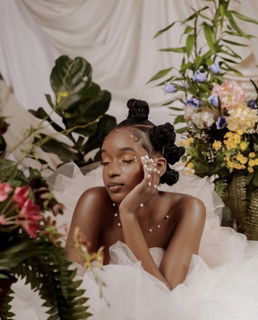 A young woman with dark skin and artistic braids, decorated with pearls and rhinestones, is resting her face on her hand among lush green and colorful flower arrangements, wearing a white, fluffy dress in a soft, elegant setting.