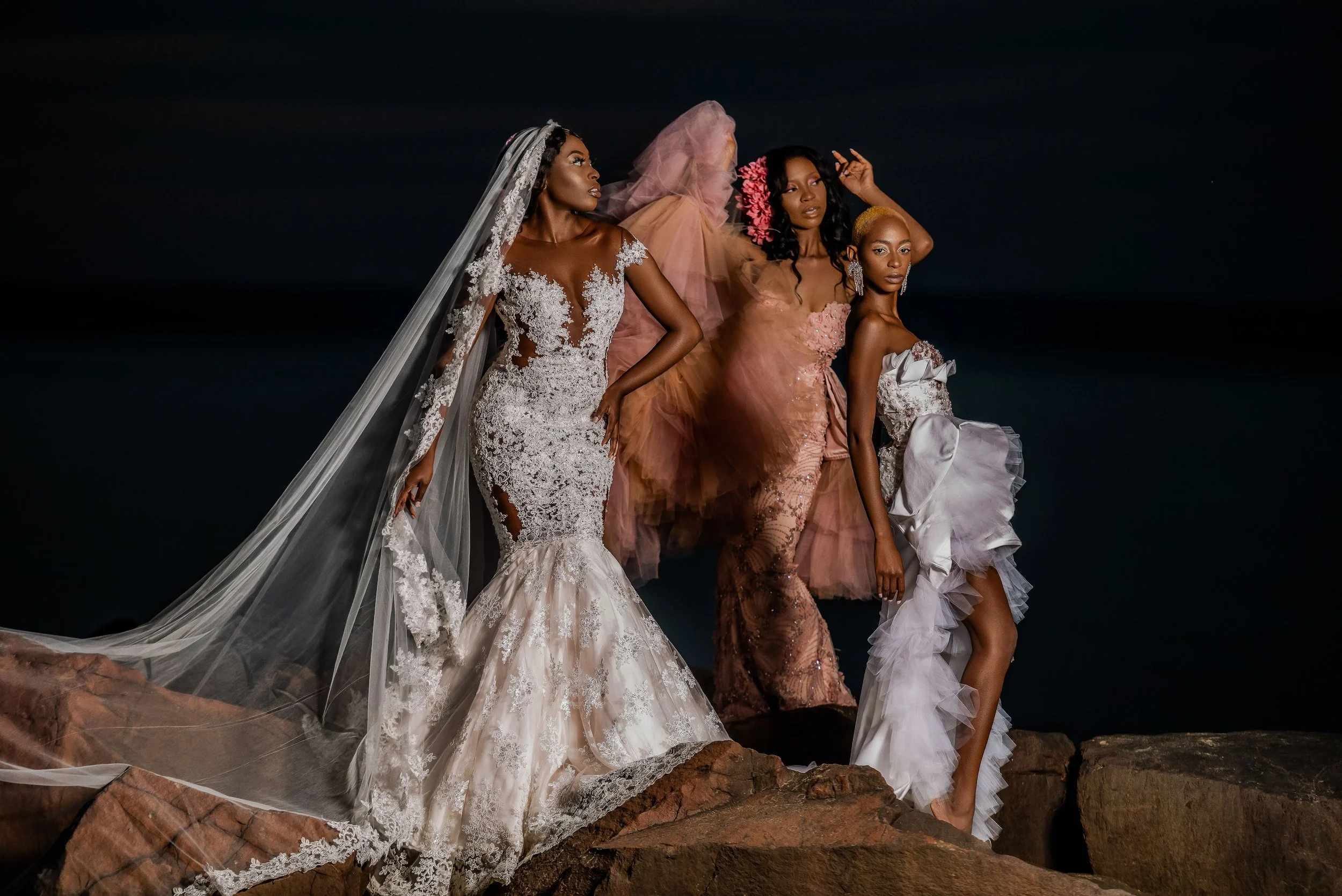 Four women wearing elaborate wedding dress gowns standing on rocks at a NJ Beach outdoors at night.