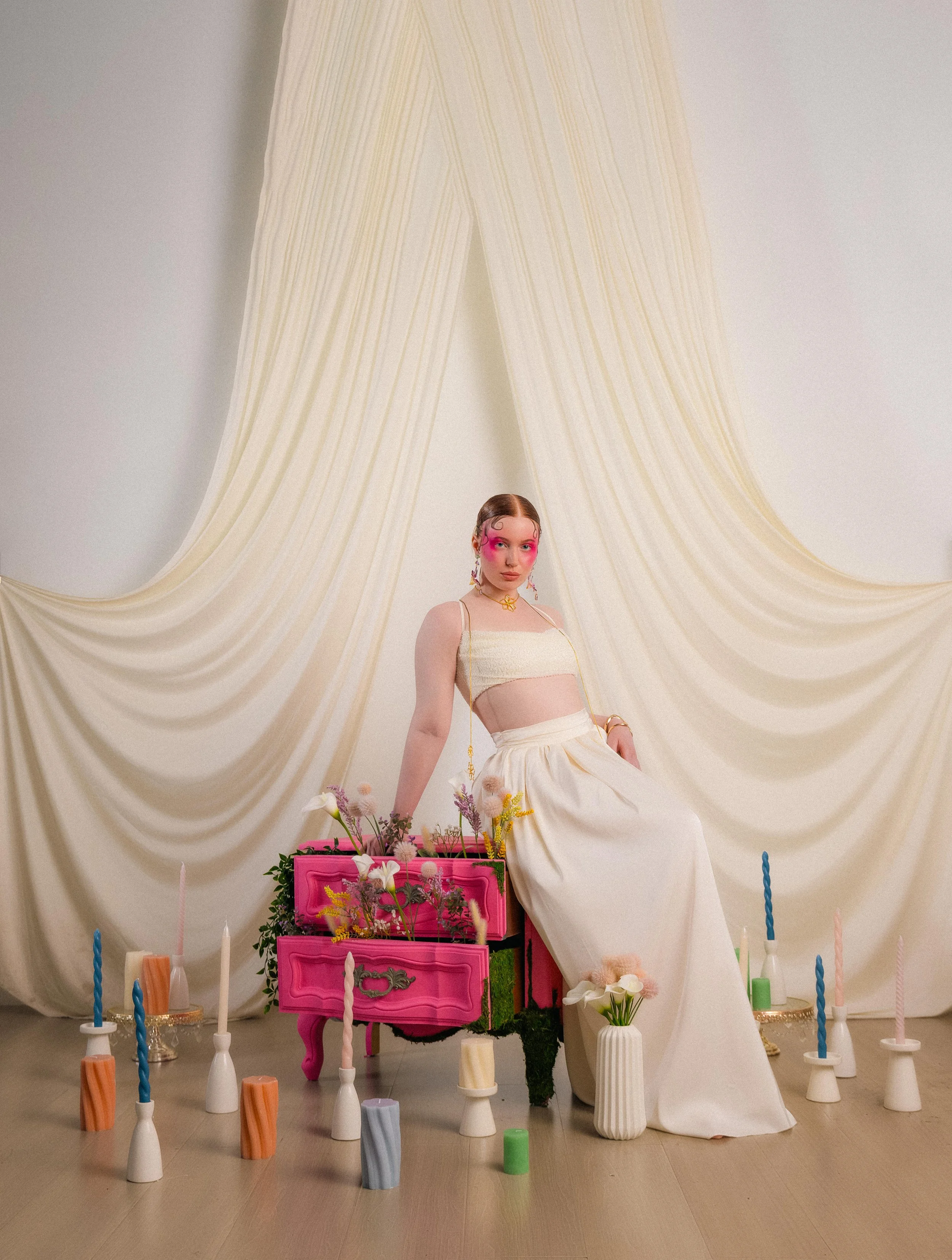 A young woman with pink makeup and colorful jewelry sitting on a pink vintage dresser decorated with flowers, surrounded by candles, in front of cream-colored draped fabric.