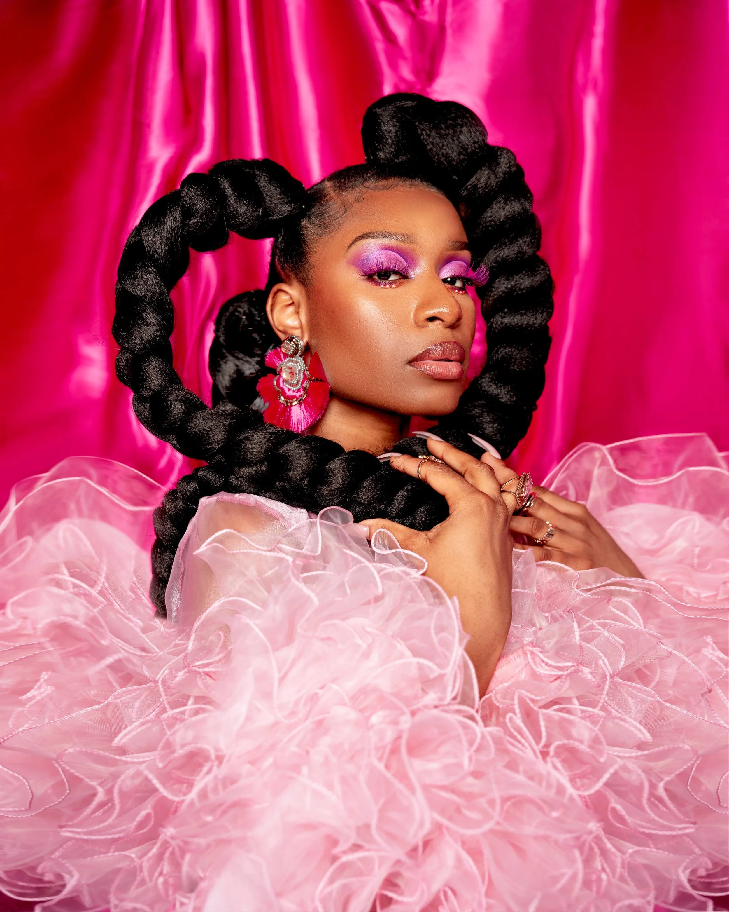 A woman with large twisted black hair styled around her head, wearing bold pink and purple makeup, large pink earrings, and a fluffy pink dress, posing in front of a pink satin backdrop.