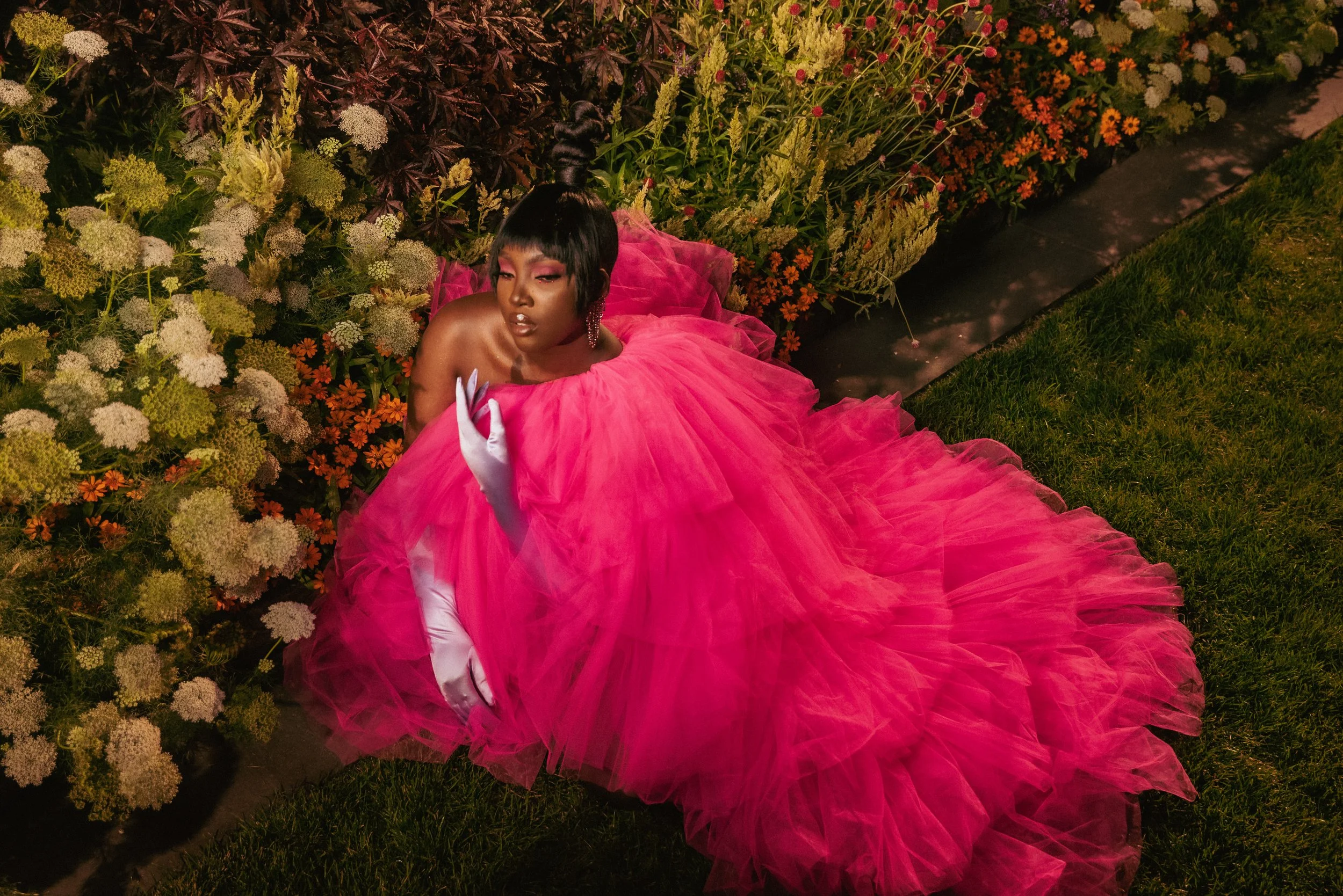A woman in a dramatic pink tulle gown with a voluminous skirt, sitting on a grassy garden path surrounded by blooming flowers.
