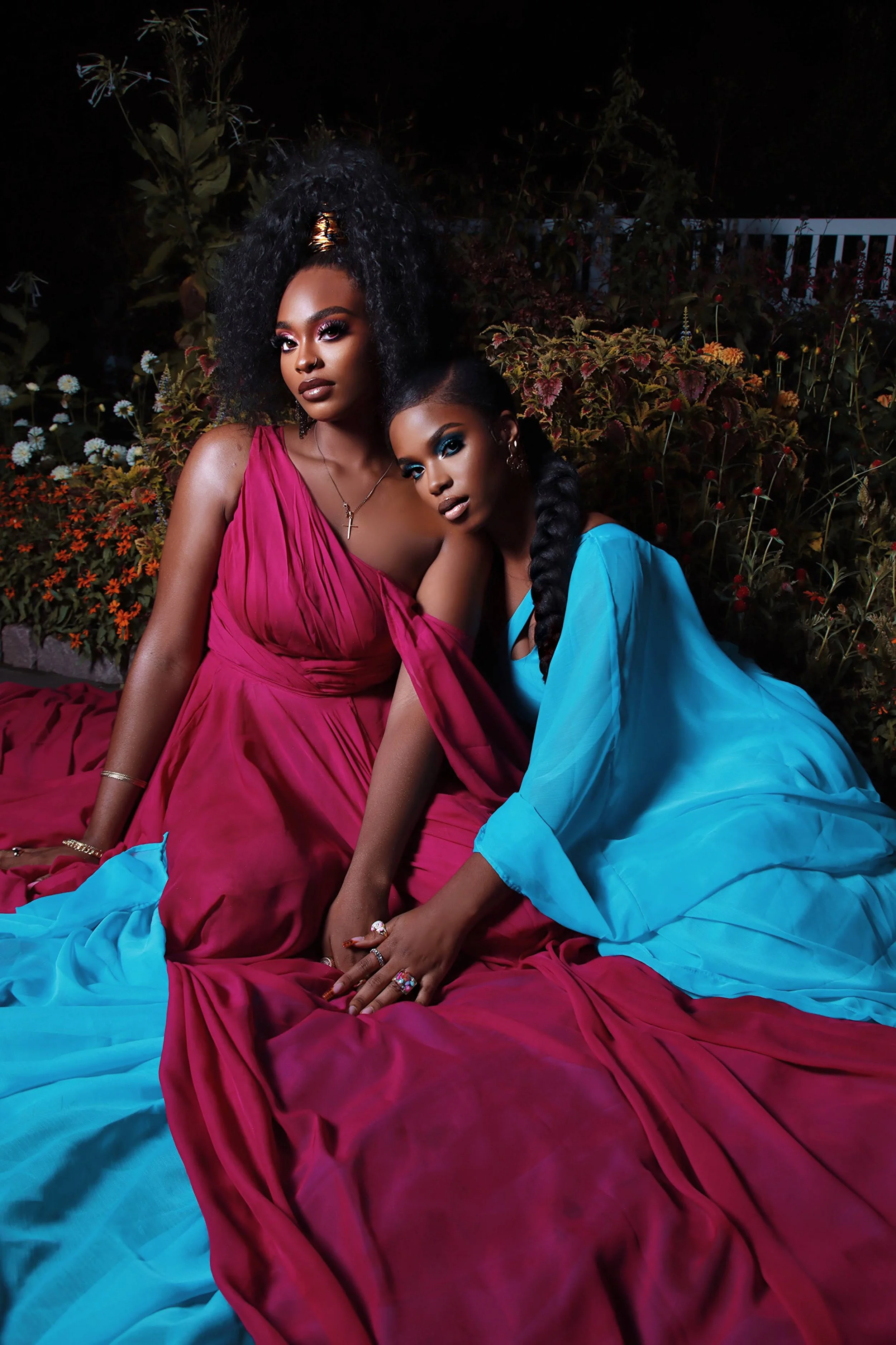 Two women wearing colorful gowns sitting close together on a bed outdoors at night, surrounded by plants and flowers.