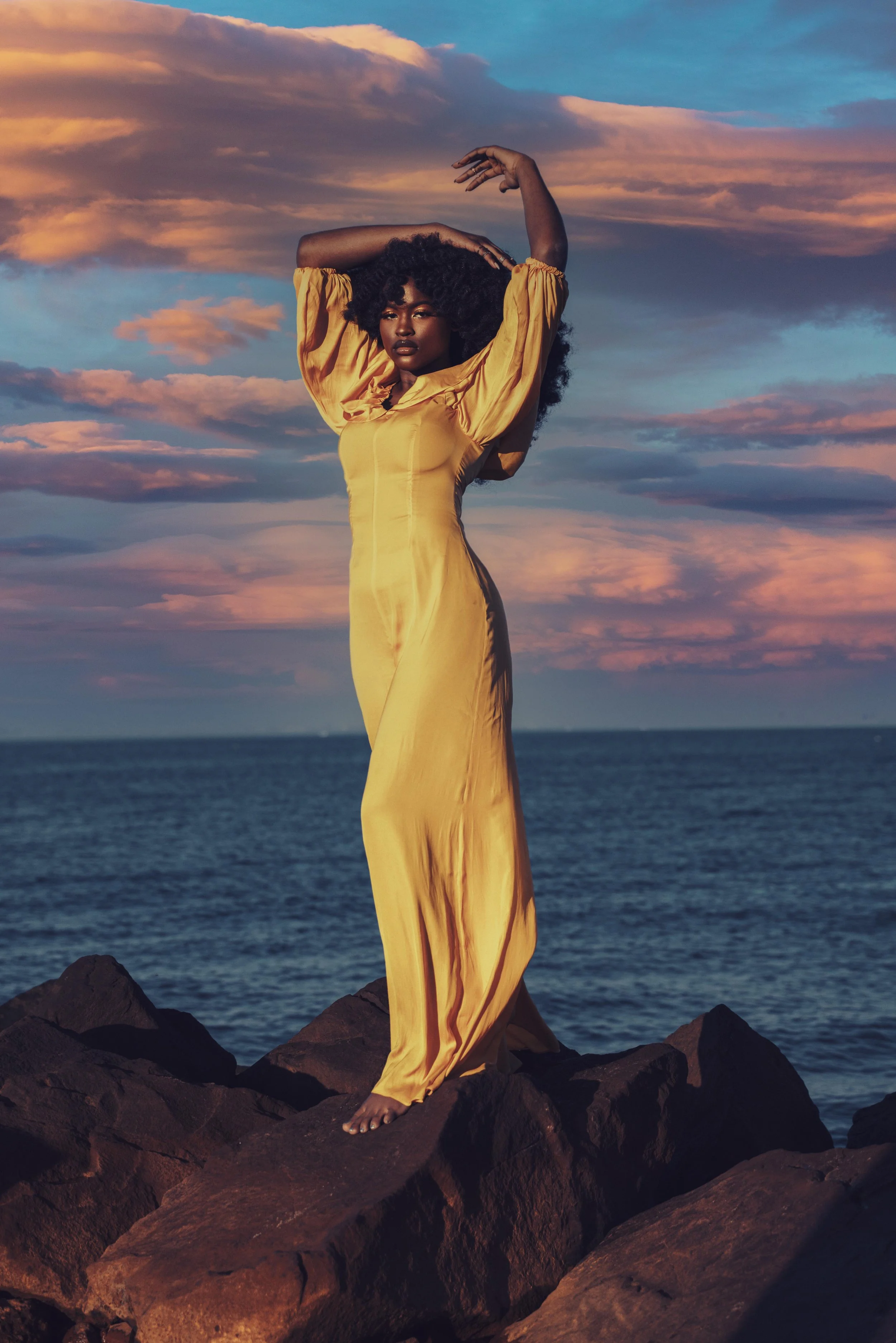 A woman in a long yellow dress stands on rocks by the ocean at sunset, with her arms raised above her head against a colorful sunset sky.