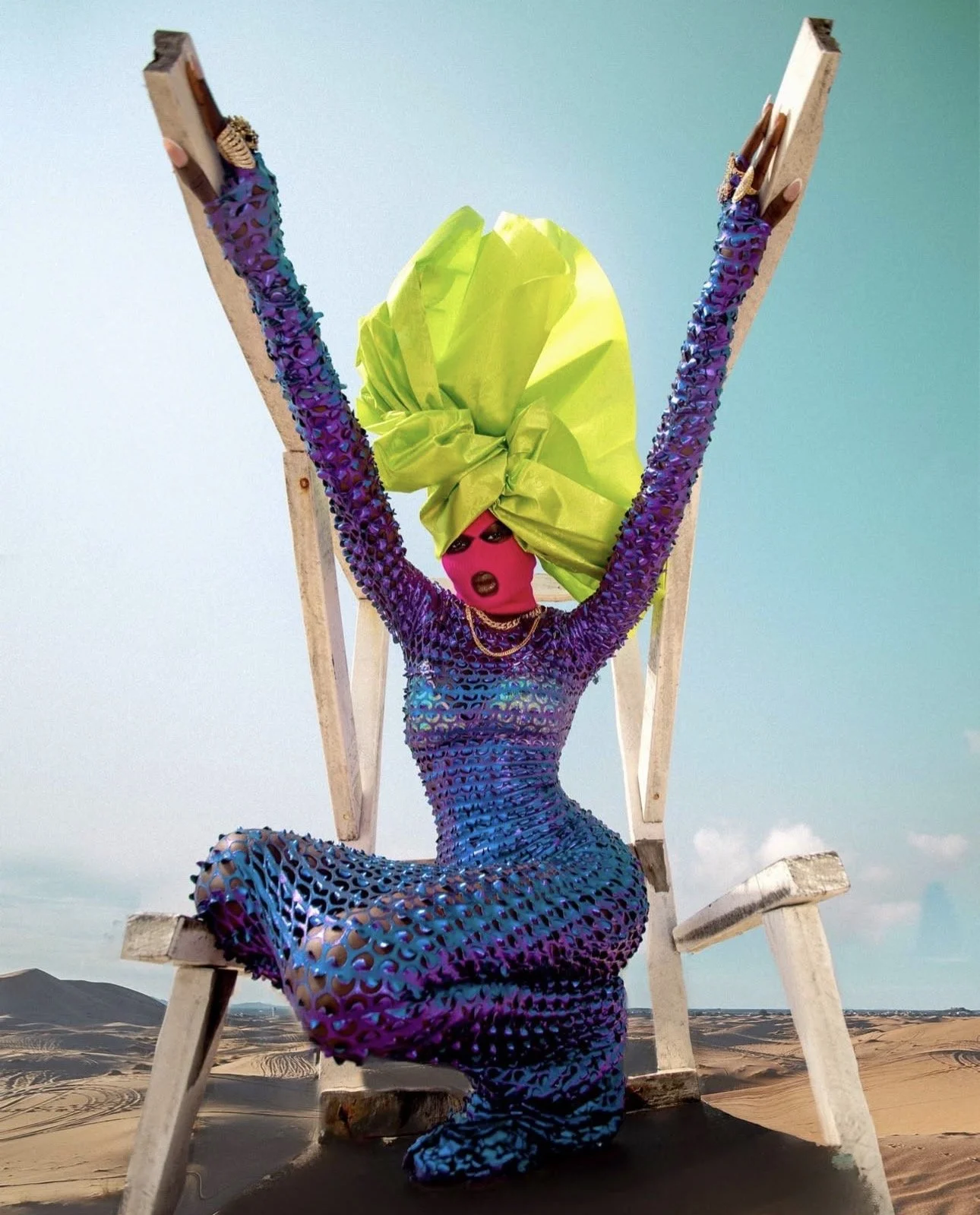 A person dressed in a vibrant, multicolored, snakeskin-like outfit with a bright yellow head wrap, wearing sunglasses and jewelry, sitting on a wooden chair in a desert landscape.