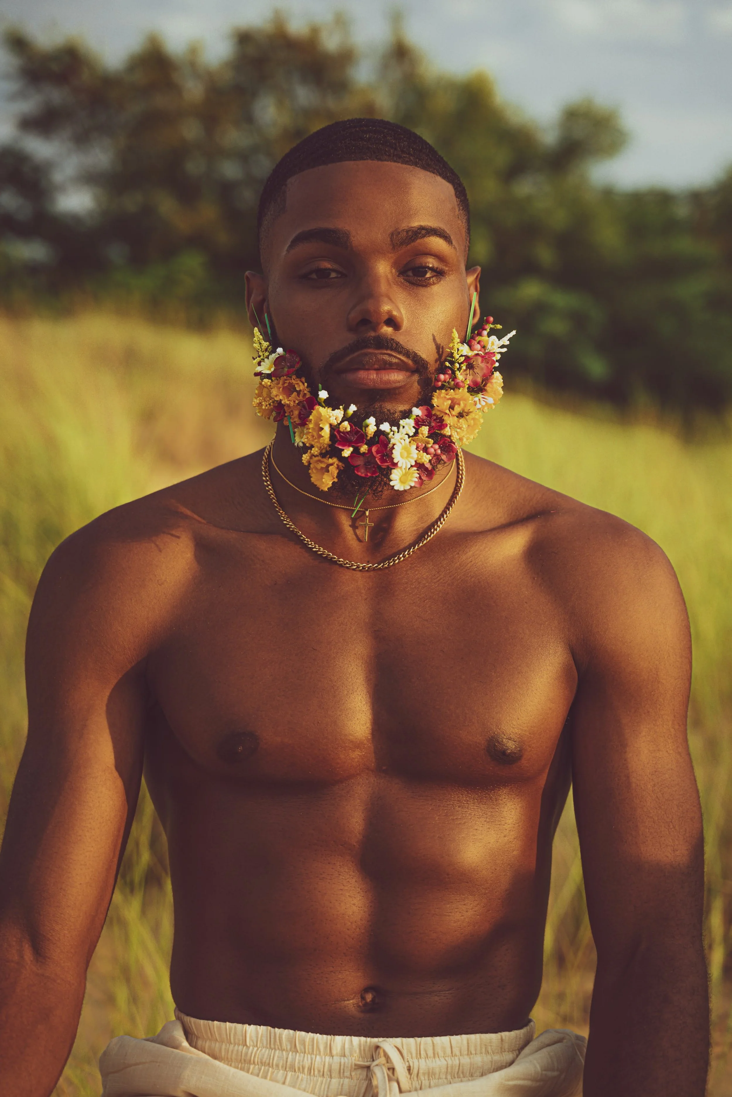 A shirtless man with a beard and short hair outdoors, wearing a floral necklace, a cross pendant, and gold chains, with a blurred natural background.