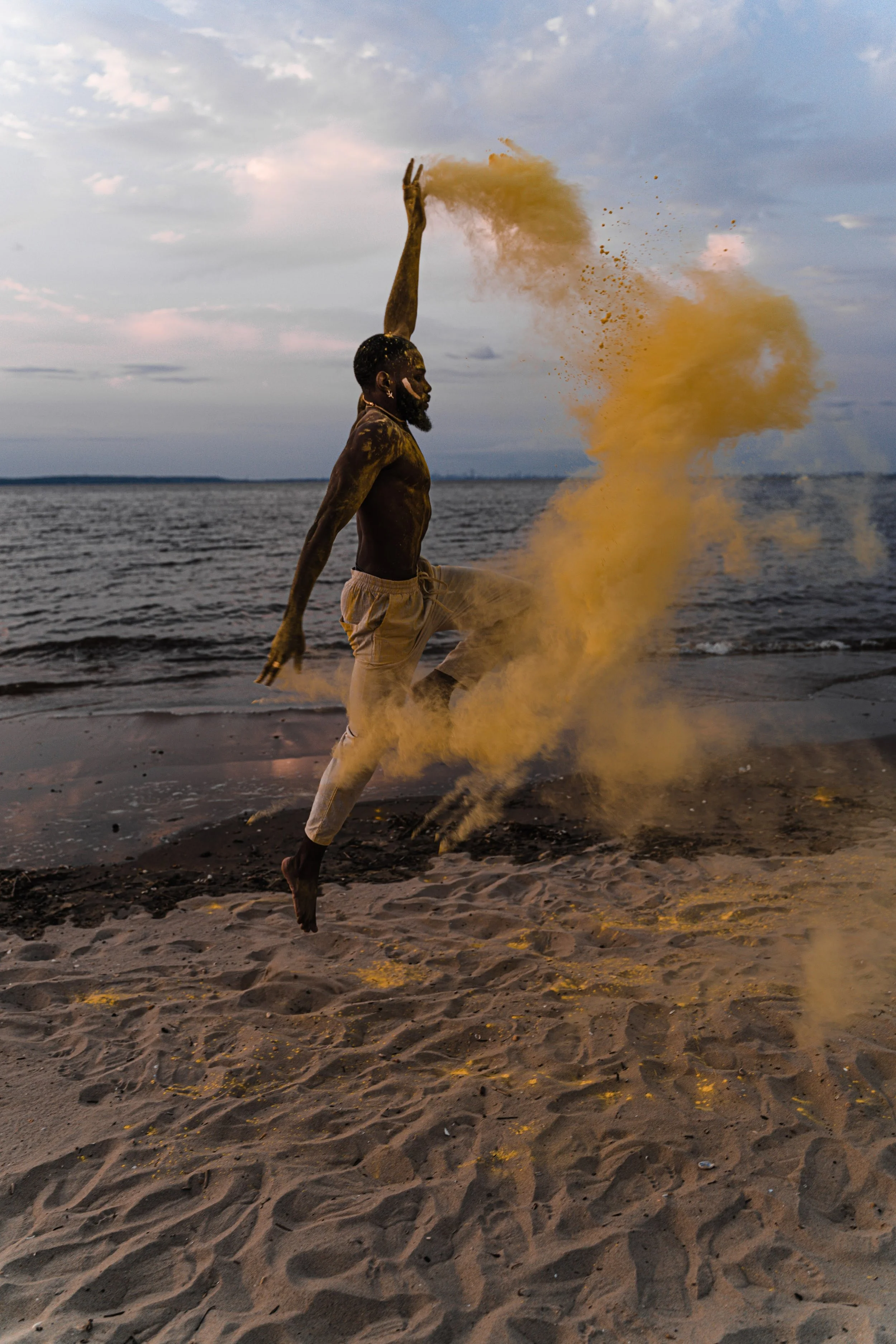 A man jumping on the beach throwing yellow powder into the air with sand and water around him during sunset or dusk.