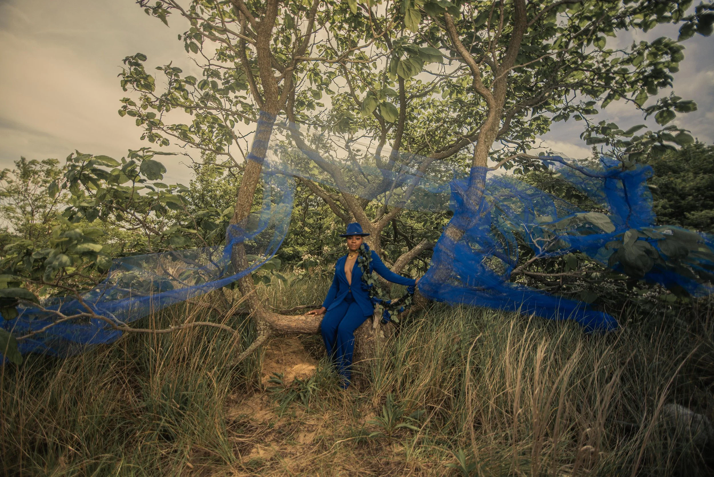 A woman dressed in a blue suit and hat sitting on a tree branch in a natural landscape, with a blue mesh fabric draped around her and the tree, and tall grass in the foreground.