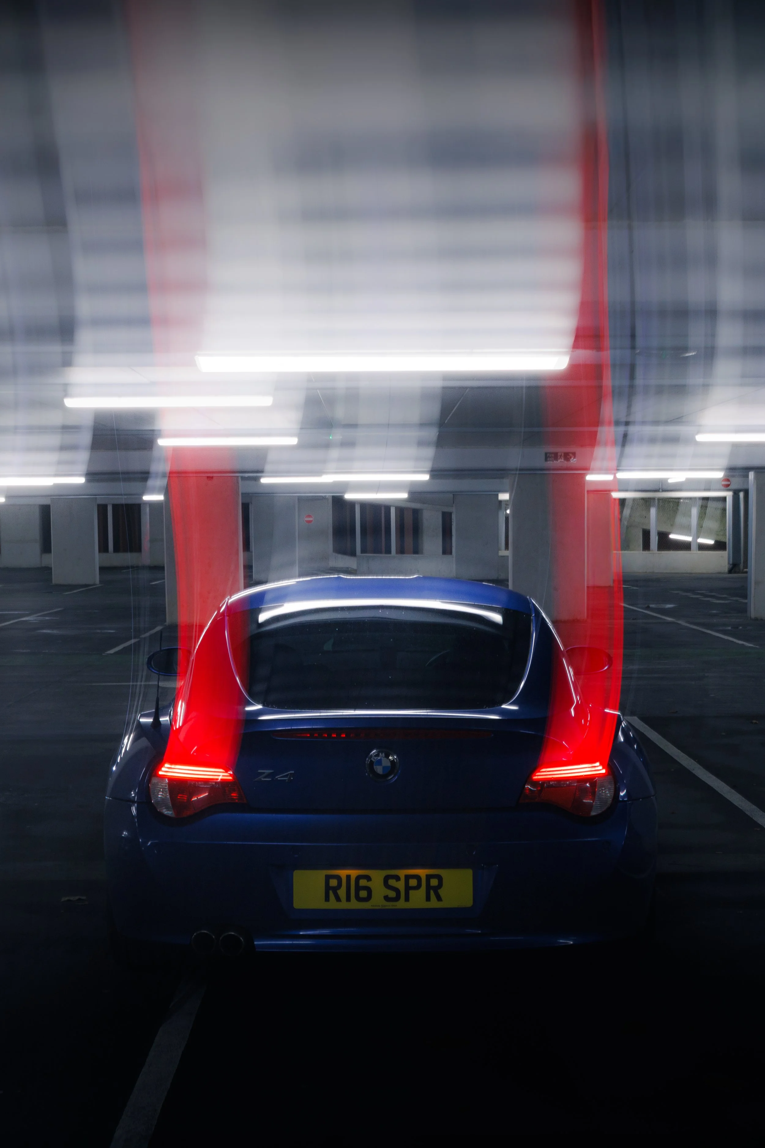 A blue BMW Z4 sports car parked in an empty parking garage at night, with light reflections on its roof and rear window, and illuminated parking lot lights overhead.