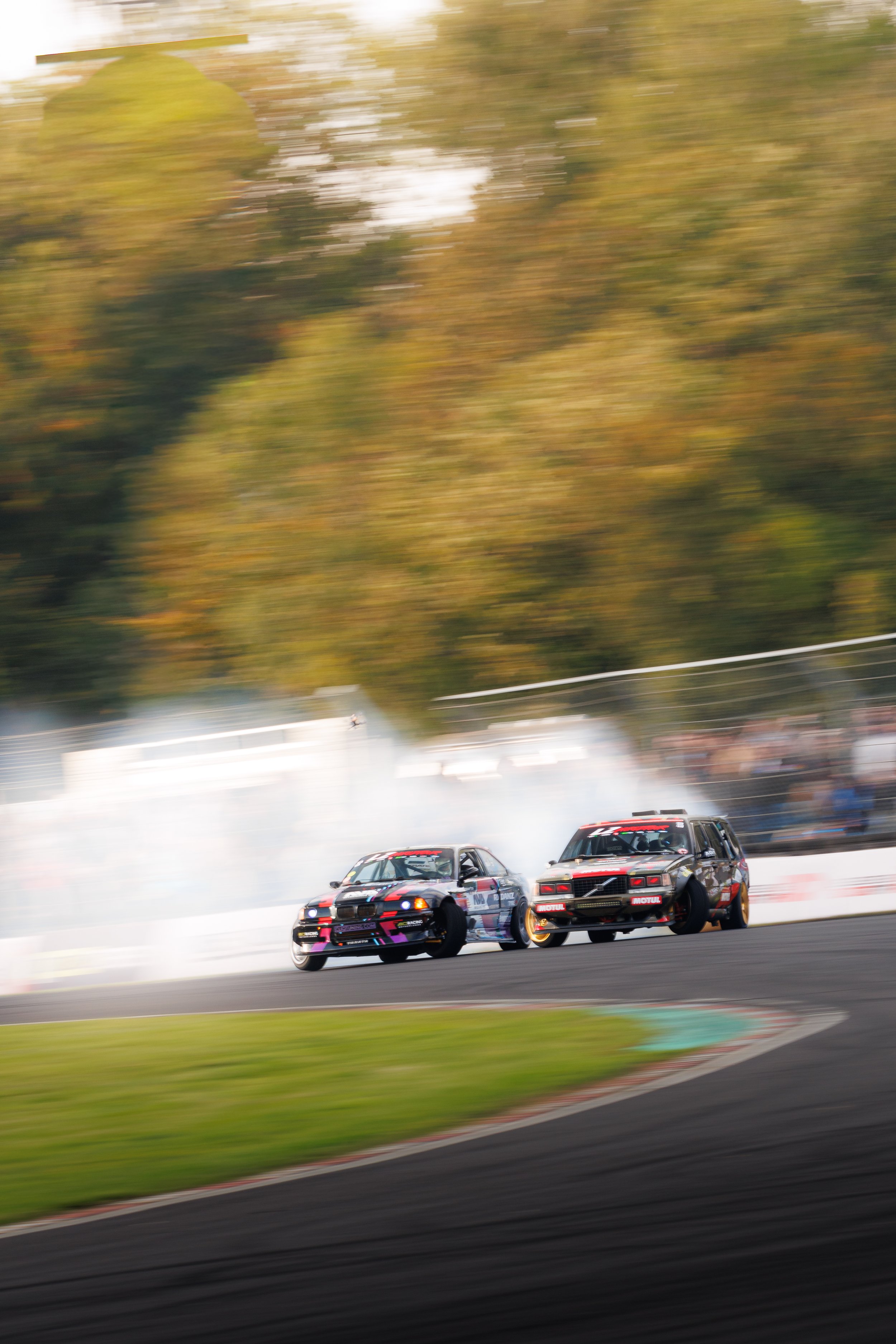 Two race cars drifting on a track, with smoke and motion blur, surrounded by blurred trees and spectators.