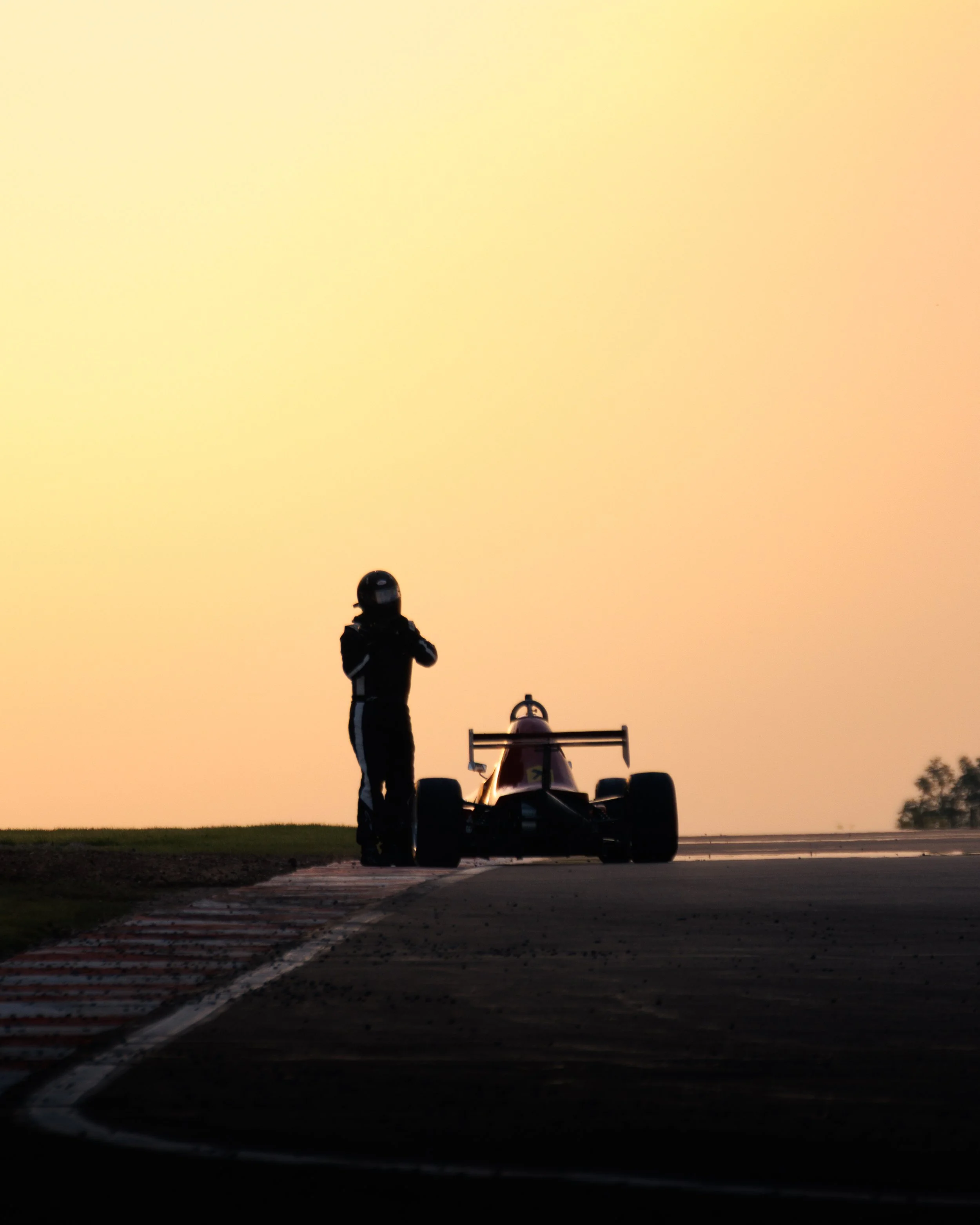 Silhouette of a race car driver standing beside a race car on a track at sunset.