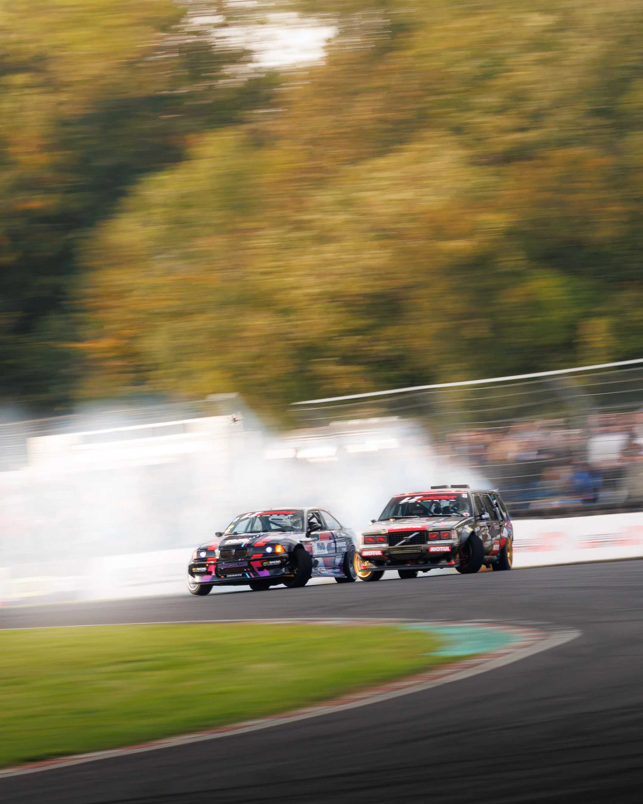 Two racing cars drifting around a track with smoke and motion blur, trees in the background.