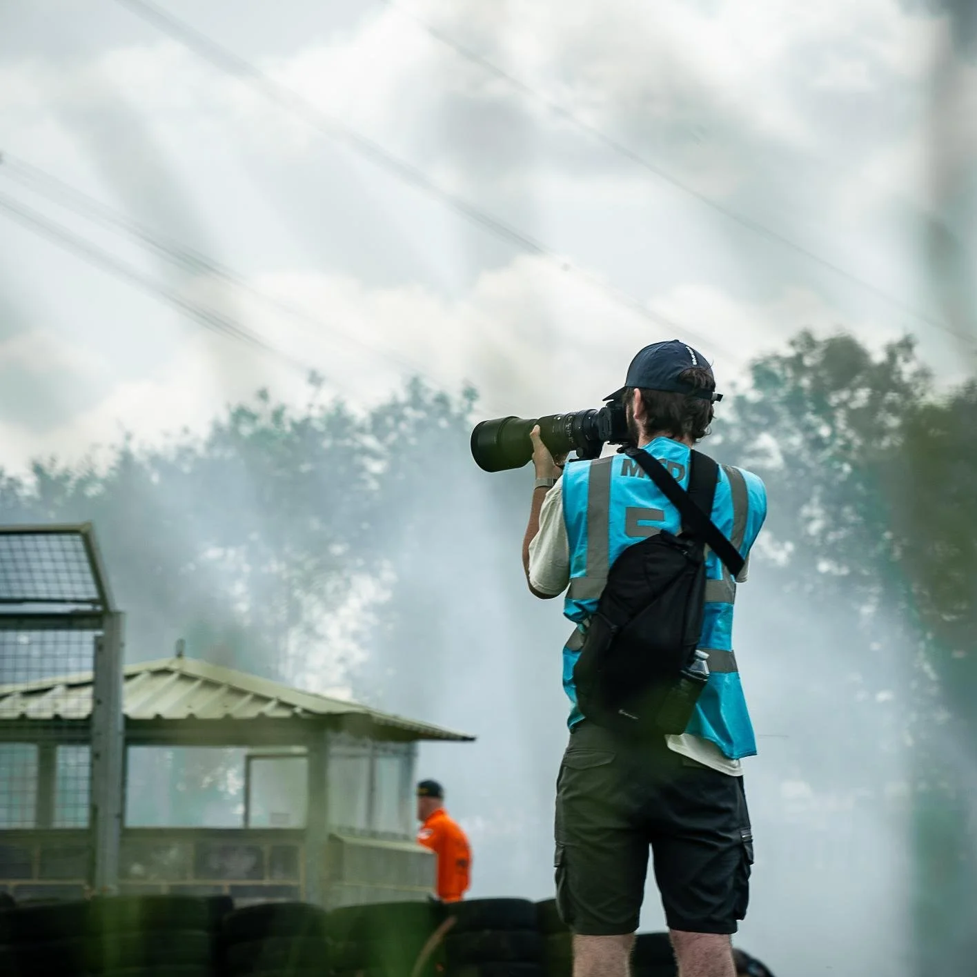 Photographer standing outdoors with a camera and telephoto lens, wearing a blue vest, black shorts, and a cap, capturing a scene at a racetrack with a person in an orange suit in the background and smoke or fog in the air.