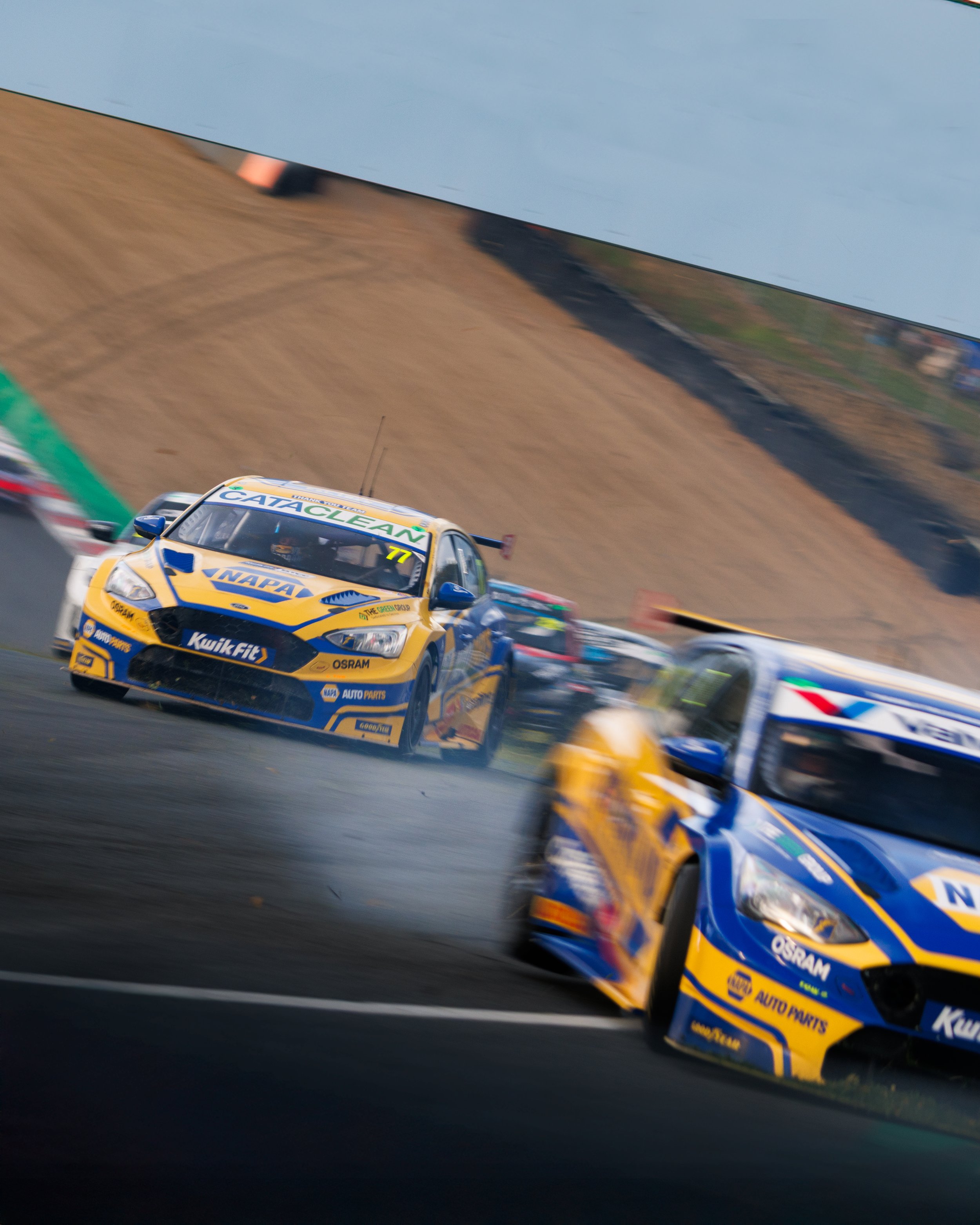 Two BTCC NAPA Racing Ford Focus cars, painted in blue and yellow with various sponsor logos, both cars are racing one of which has lost control and is moving towards the camera
