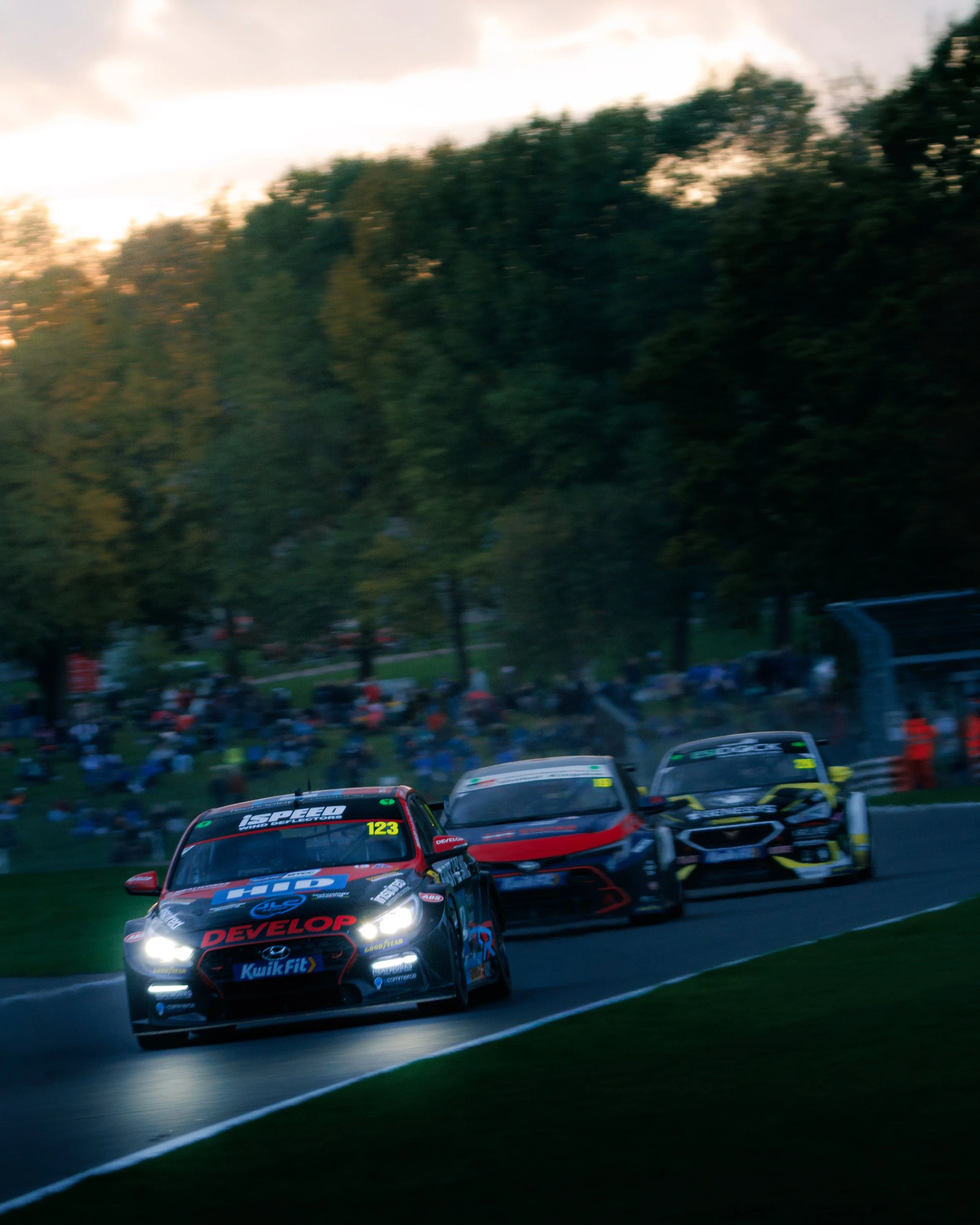 Three race cars on a racetrack during dusk, cornering with blurred spectators and trees in the background.