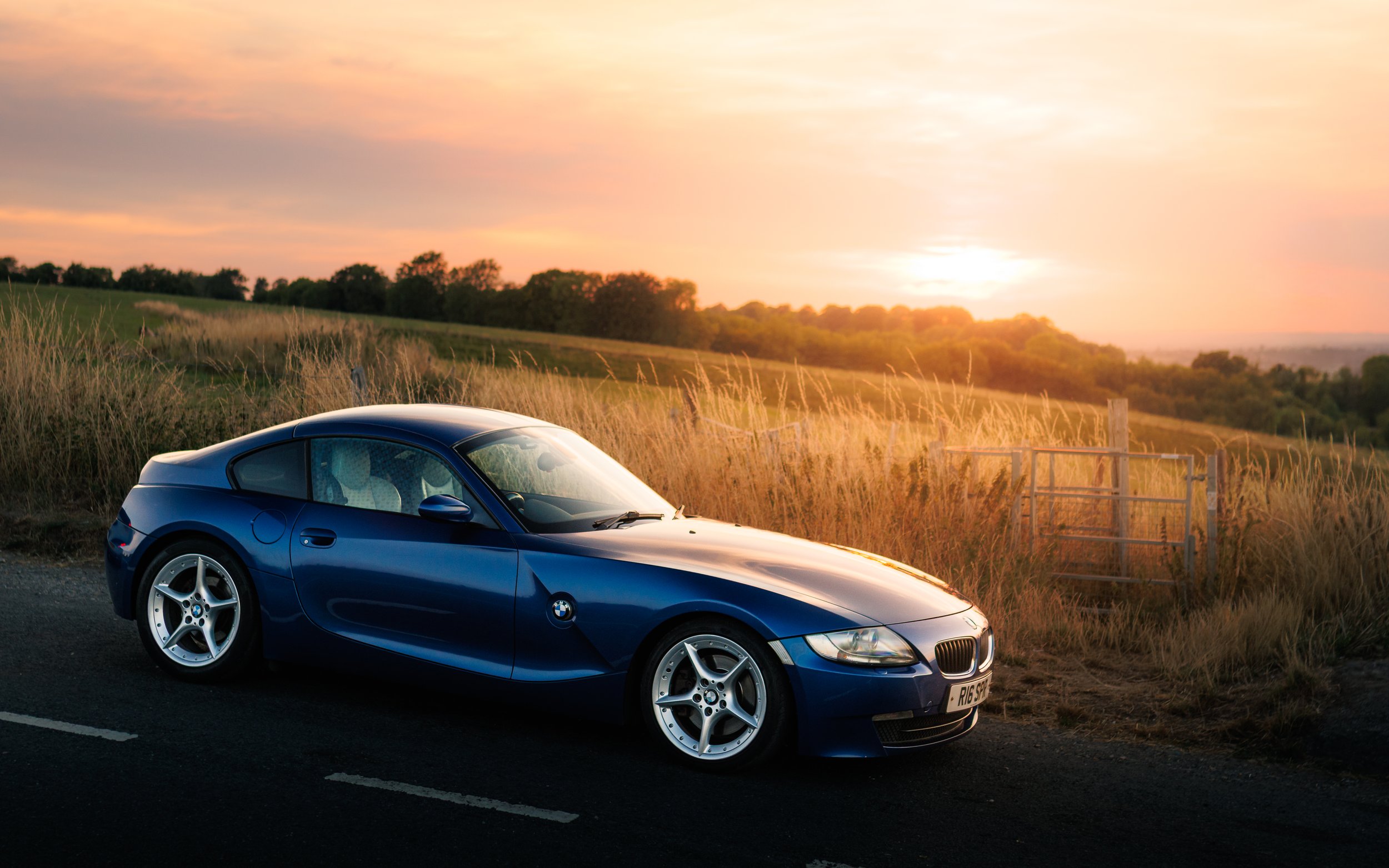A blue BMW sports car parked on the side of a rural road during sunset, with tall grass and a field in the background.