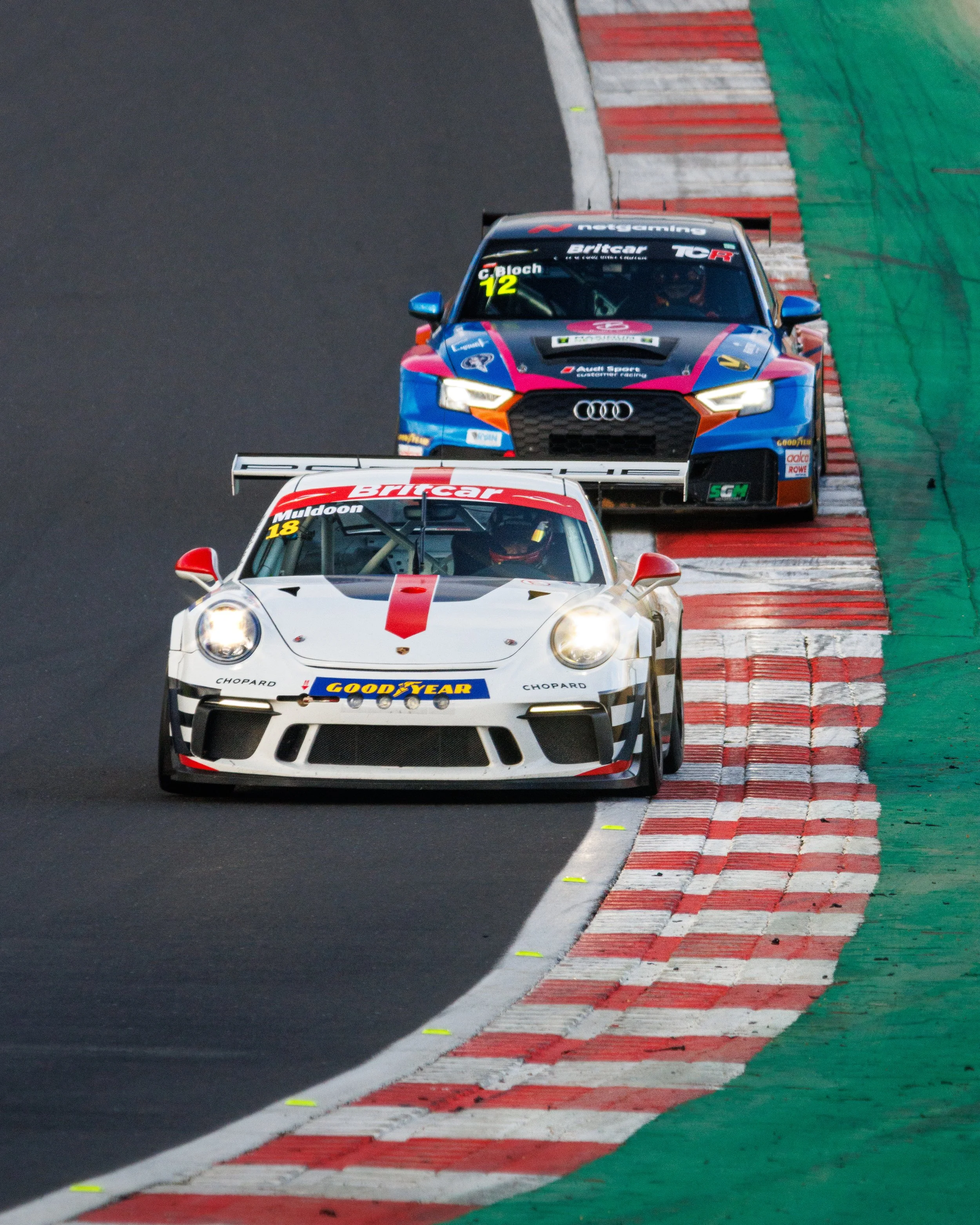 Two race cars on a track, one leading and the other following closely behind, as they navigate a corner.