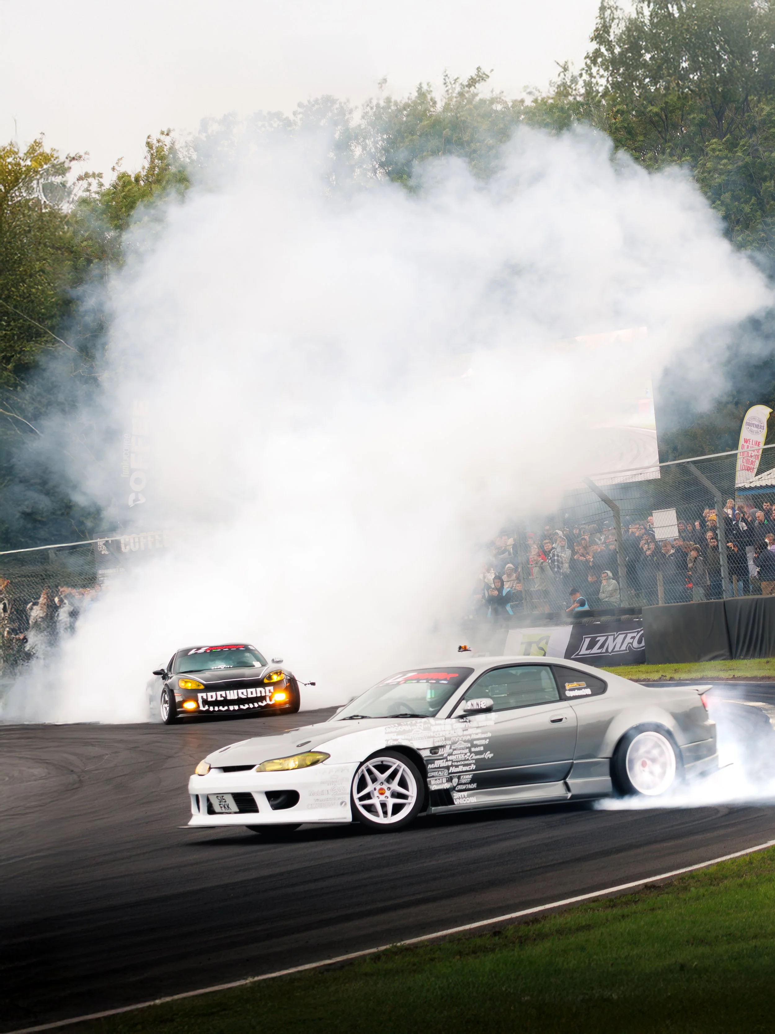 Two cars drifting on a race track, with smoke billowing behind them and spectators in the background.