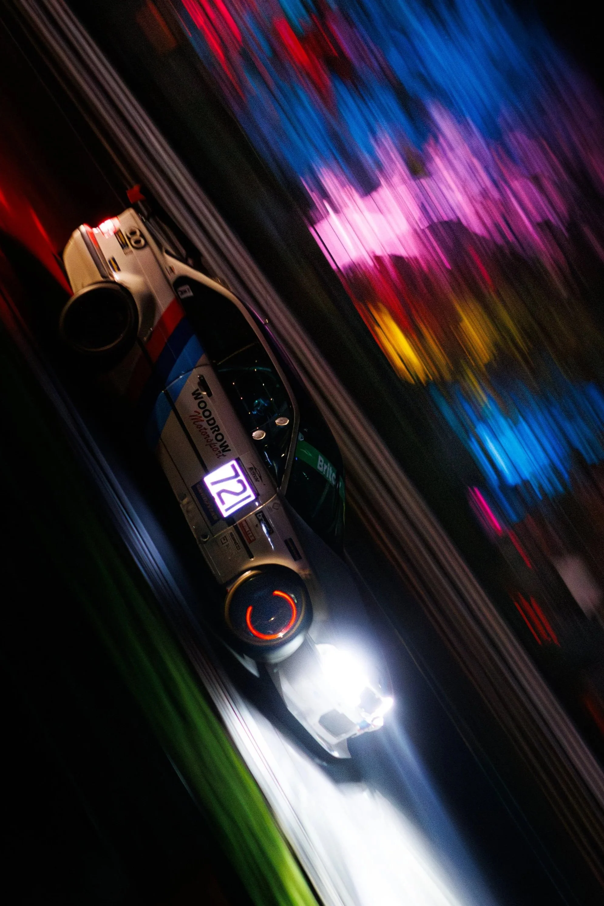 Photo of a race car speeding on a track at night, with glowing brakes and colourful blurred lights in the background.