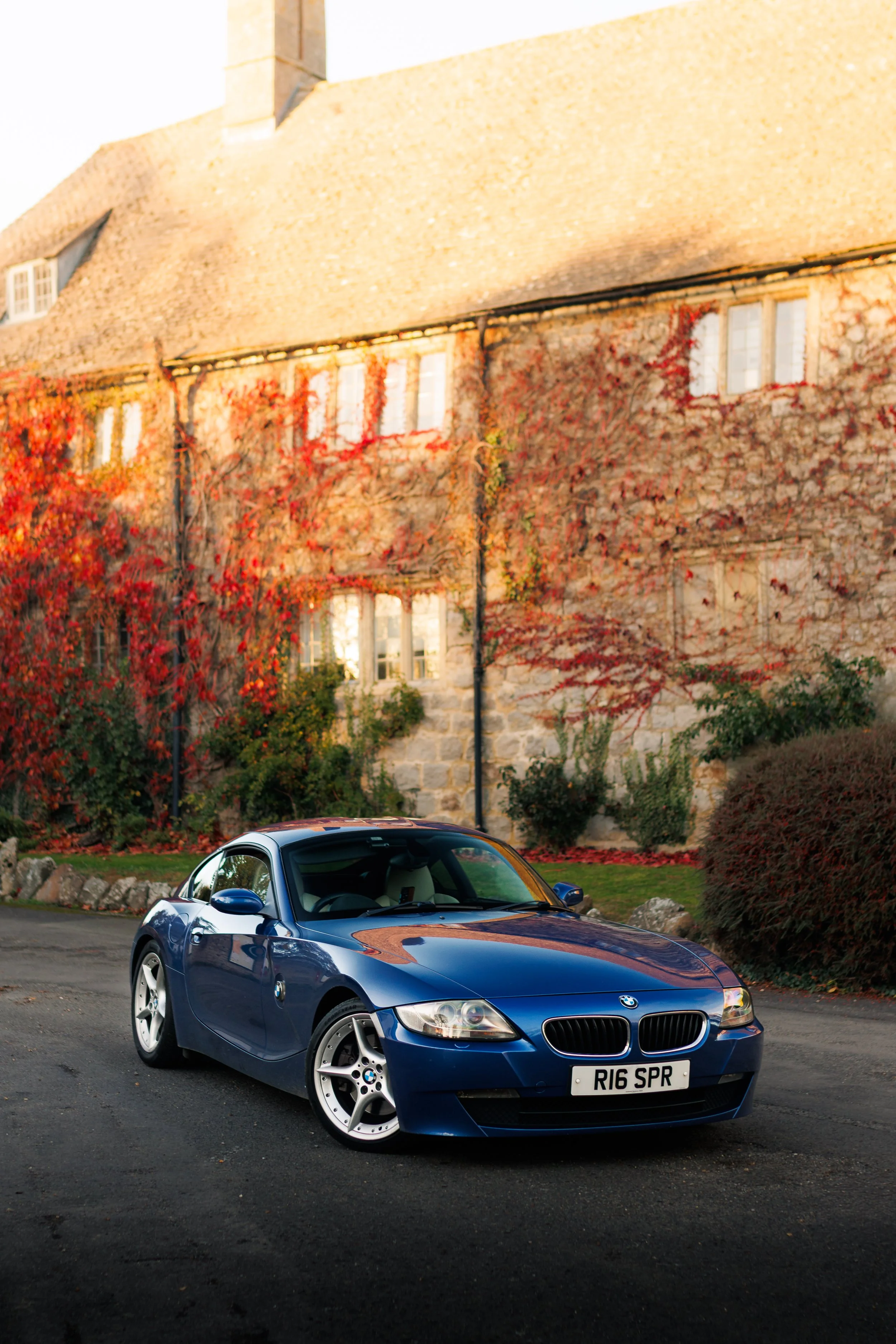 A blue BMW Z4 sports car parked on a driveway in front of a stone house with ivy-covered walls and autumn foliage.