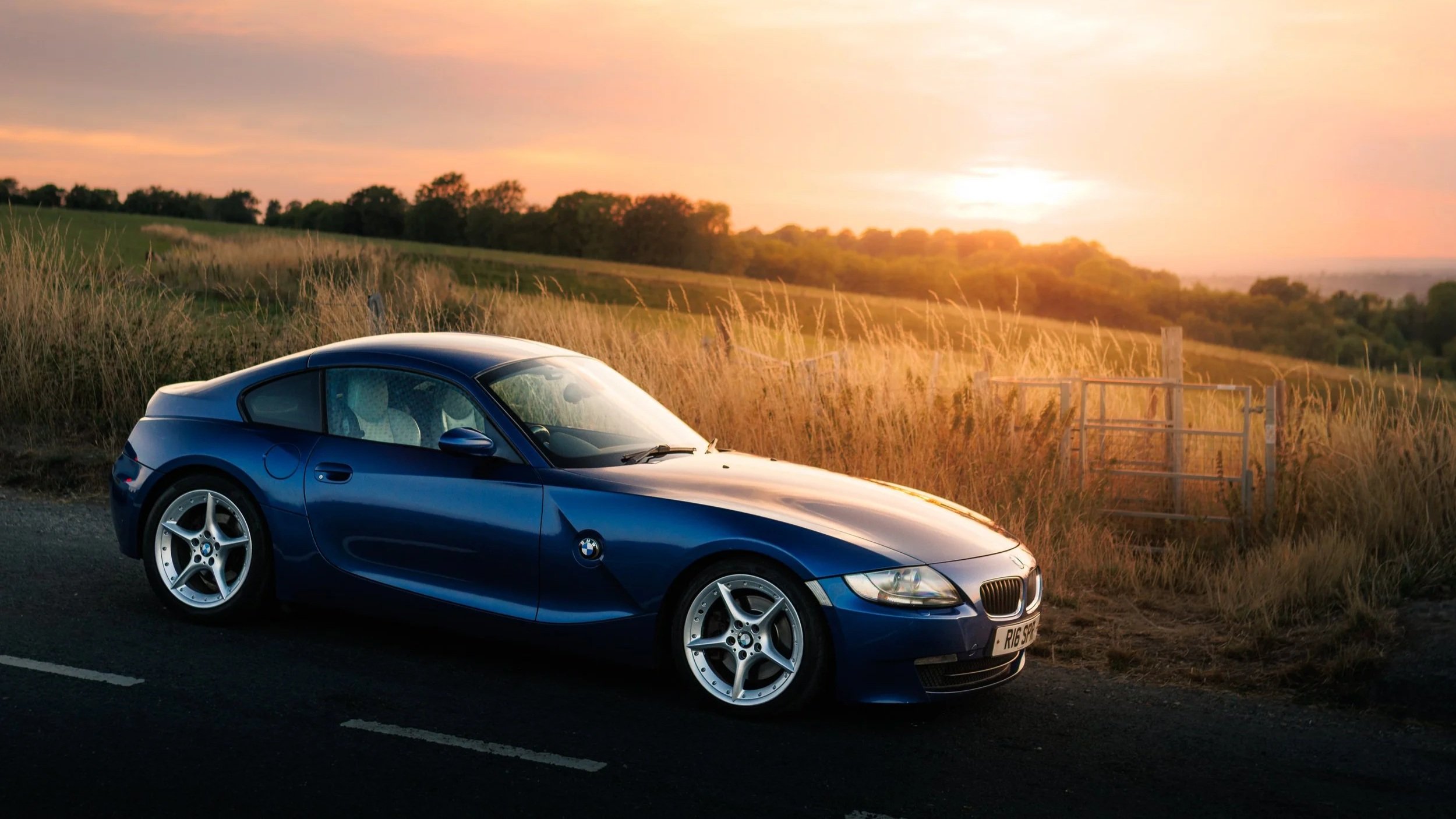 A blue BMW sports car parked on the side of a rural road during sunset with a grassy field and trees in the background.