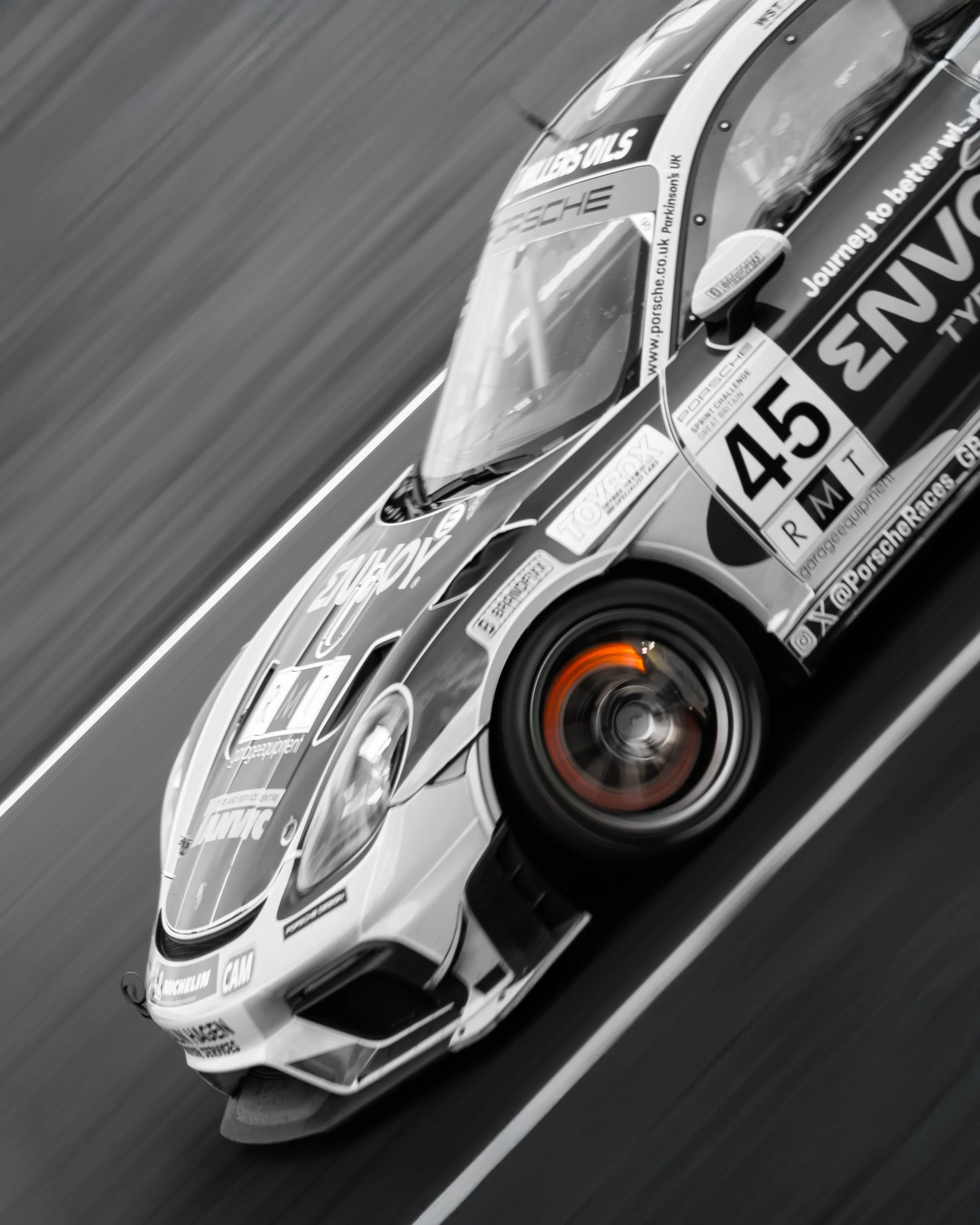 Black and white photo of a Porsche race car with it's glowing brake remaining in colour moving on a racetrack at high speed, with visible sponsor logos and a blurred background.