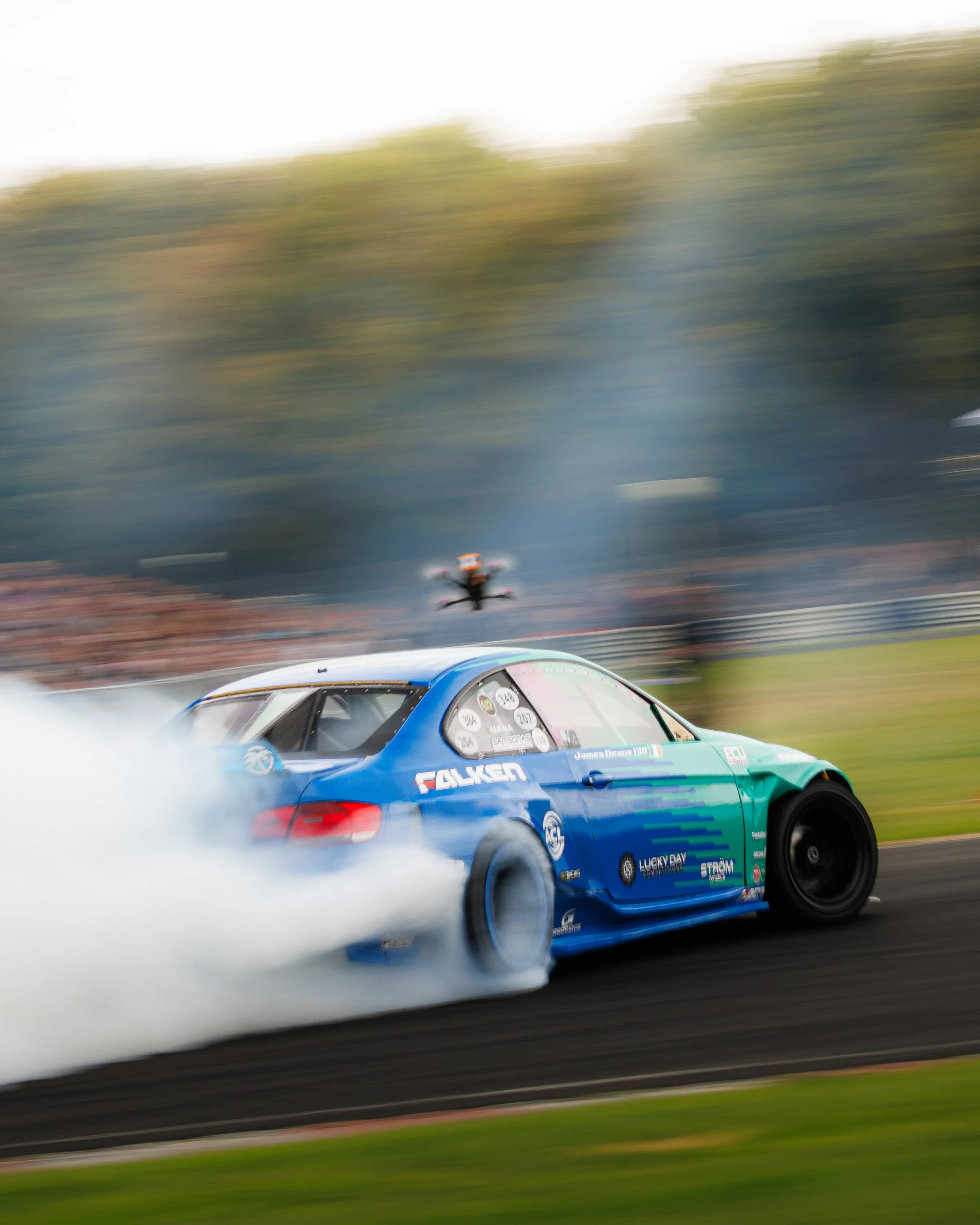 A blue race car drifting on a track, producing tire smoke, during a motorsport event with a blurred background of trees and a drone flying overhead.