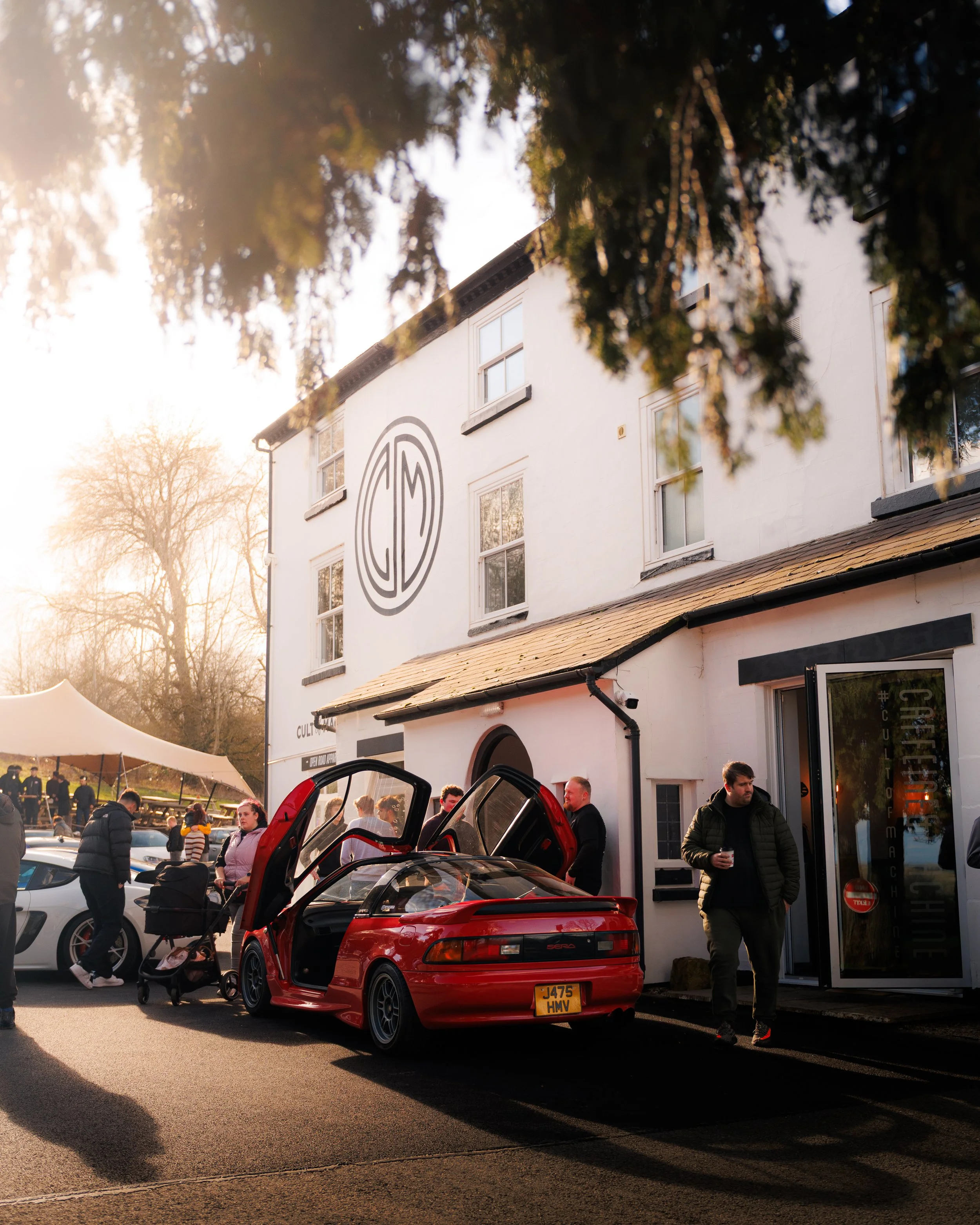 A group of people gathering outside Caffeine and Machine, a white building with a logo on the wall, a red sports car with its gullwing doors open, and a man walking past holding a drink, during sunset.
