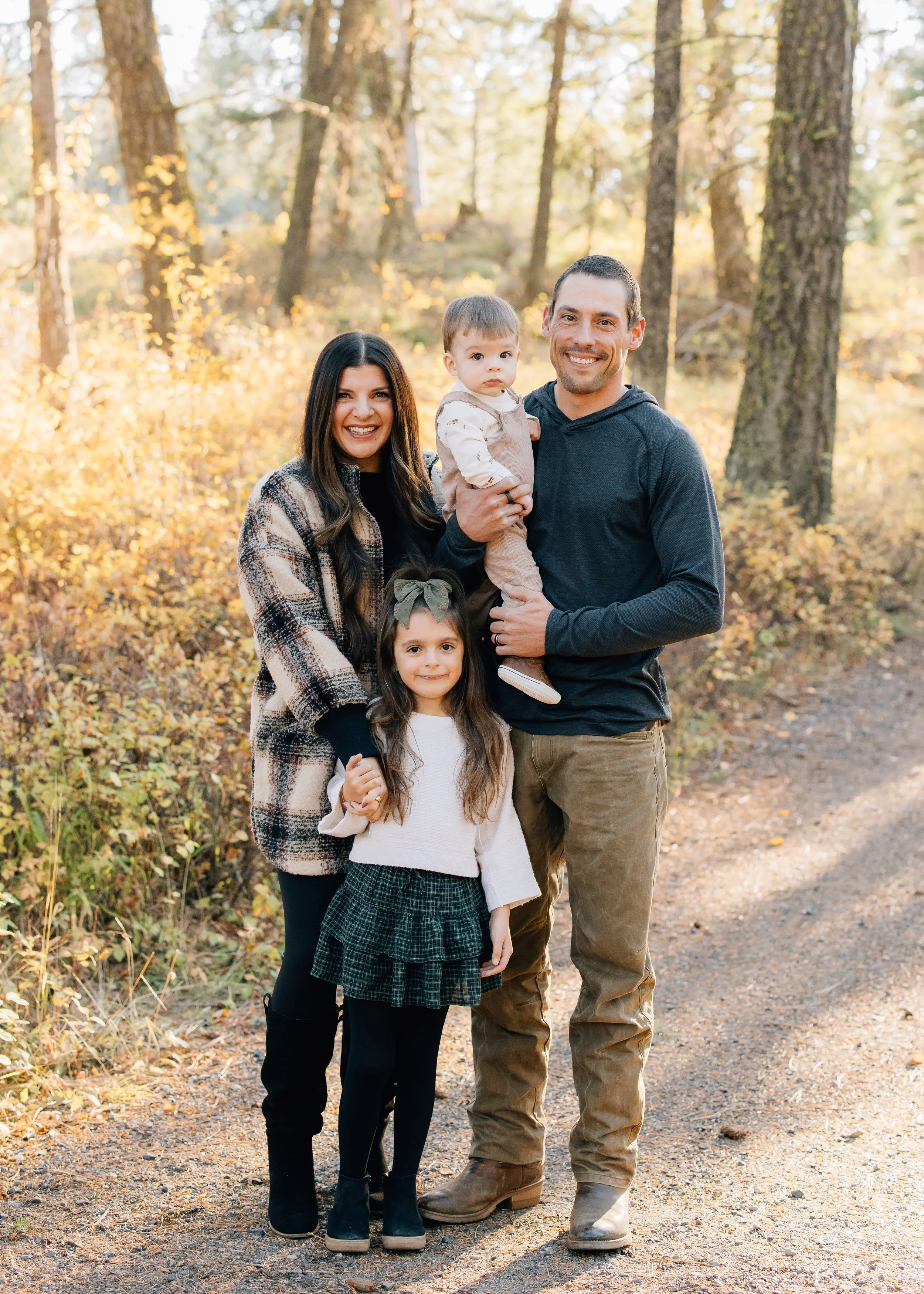 A family of four posing outdoors in a forest during autumn. The mother, father, daughter, and son are smiling and holding hands.