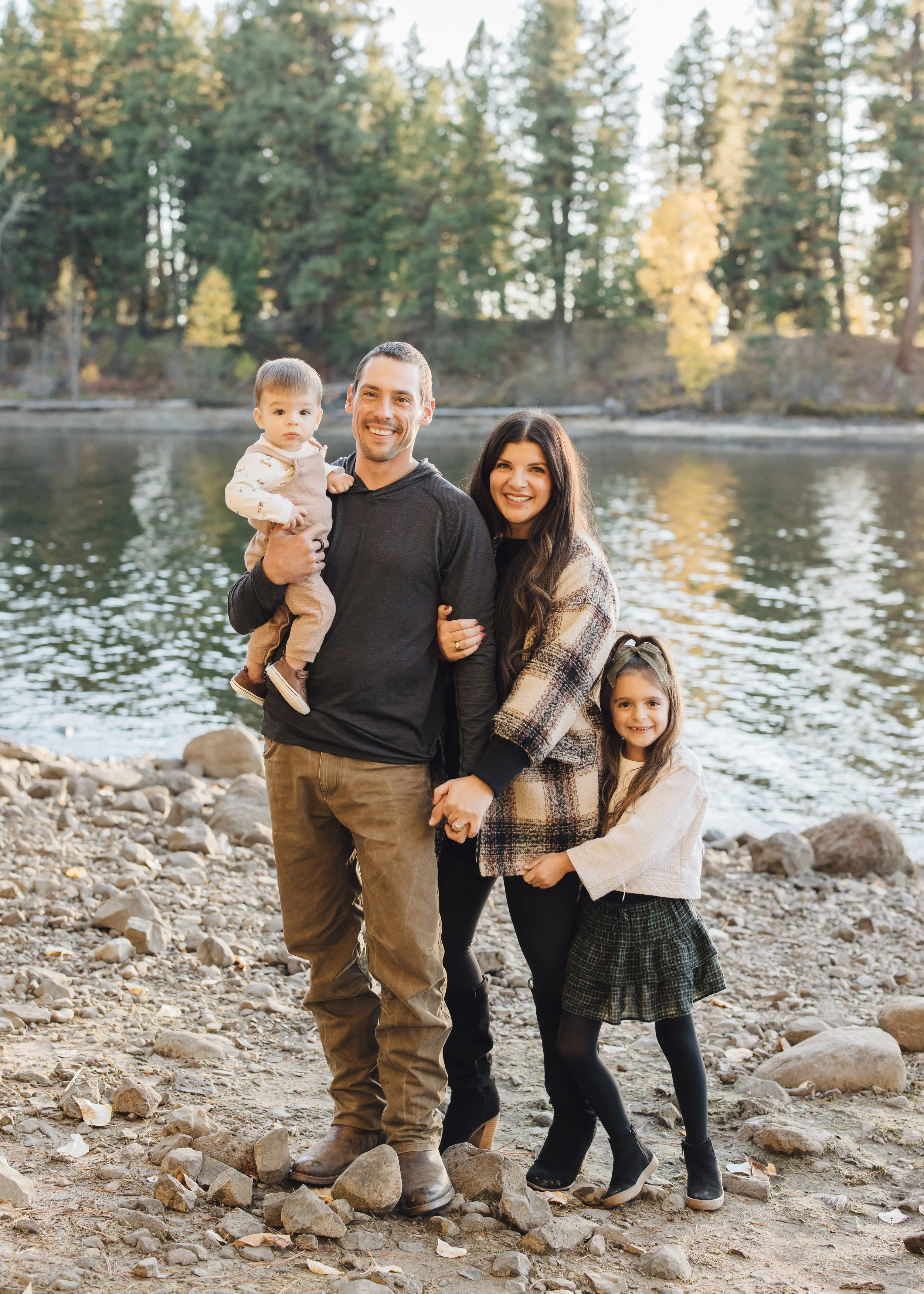 A family of four standing on a rocky shoreline by a lake, with trees in the background and autumn foliage. The father is holding a young boy, the mother is holding hands with a young girl, all smiling at the camera.