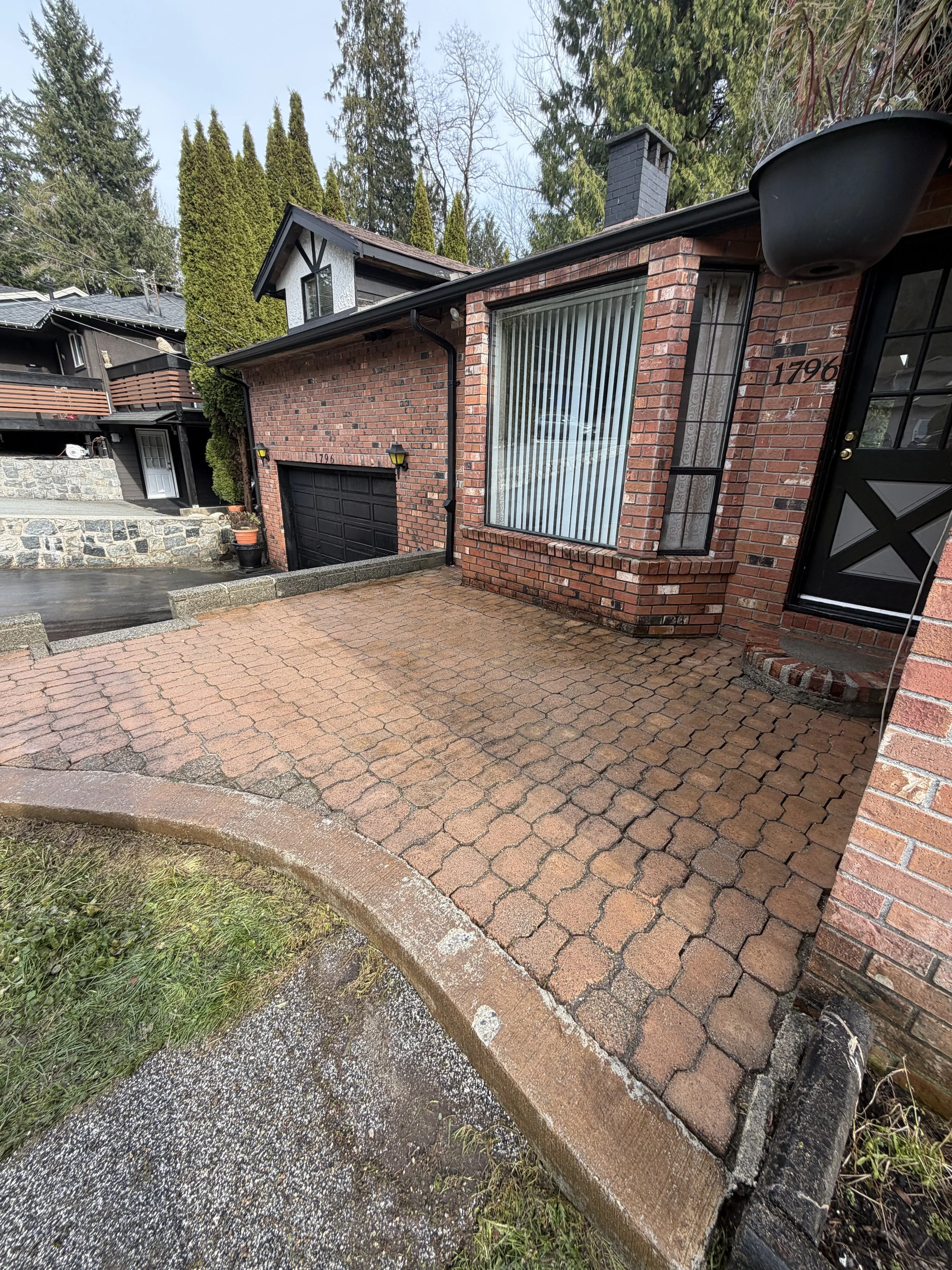 Brick house with a large window with vertical blinds, black front door, and a two-car garage with black doors. The driveway is made of red interlocking bricks, and there are tall trees and other houses in the background.