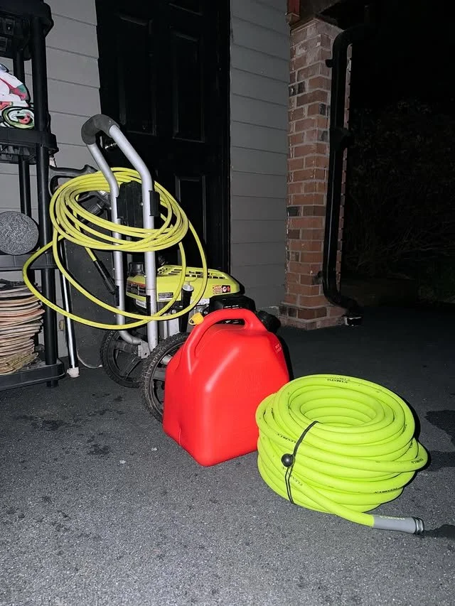 A yellow garden hose is coiled on the ground and another yellow hose is hanging on a cart. There is a red gasoline container and a small portable pressure washer with a yellow body and black wheels. The scene is outside a house with gray siding and a brick column, next to a black door.