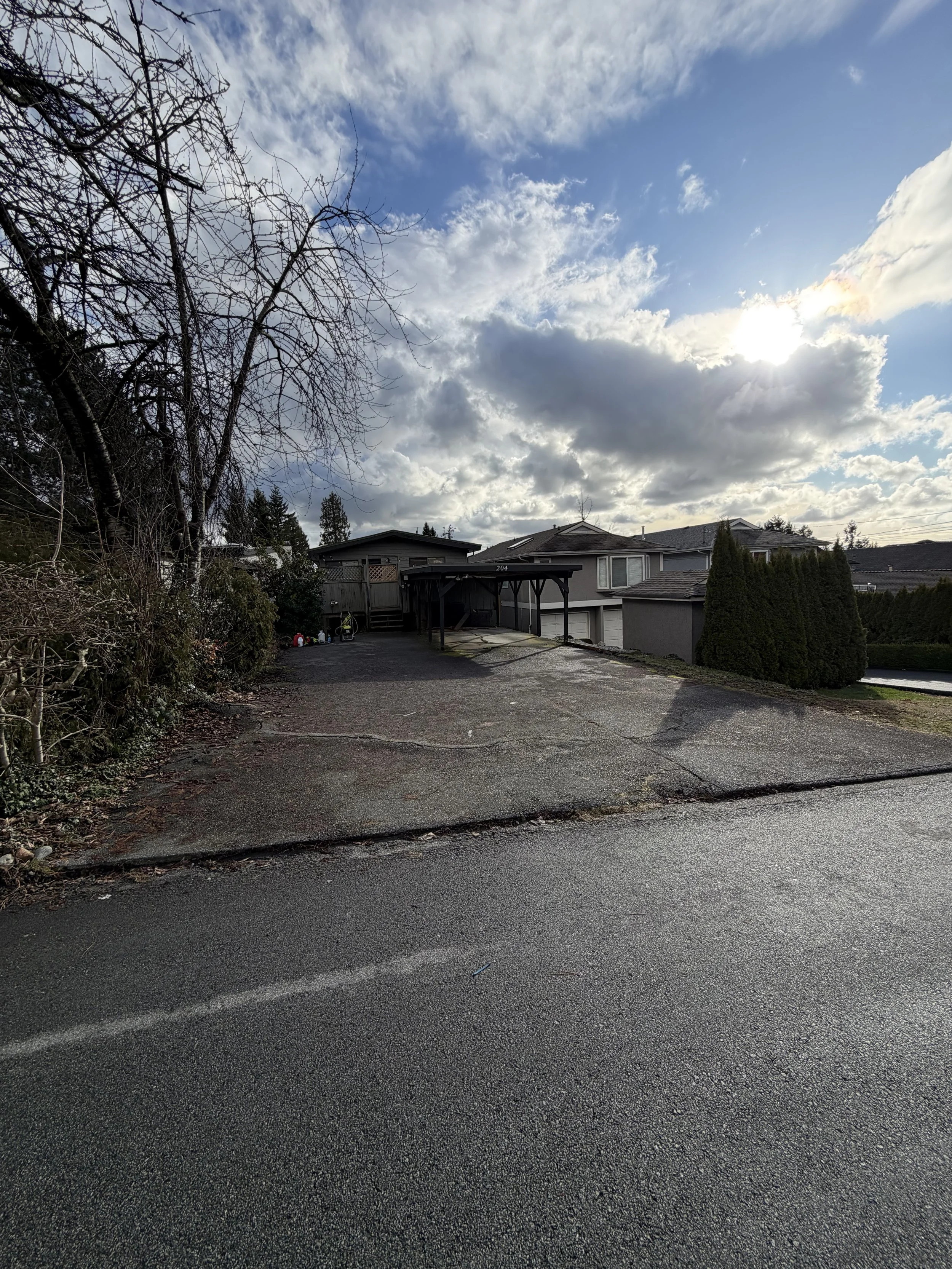 View of a residential driveway and house with a carport, surrounded by trees, with a cloudy sky and sunlight in the background.