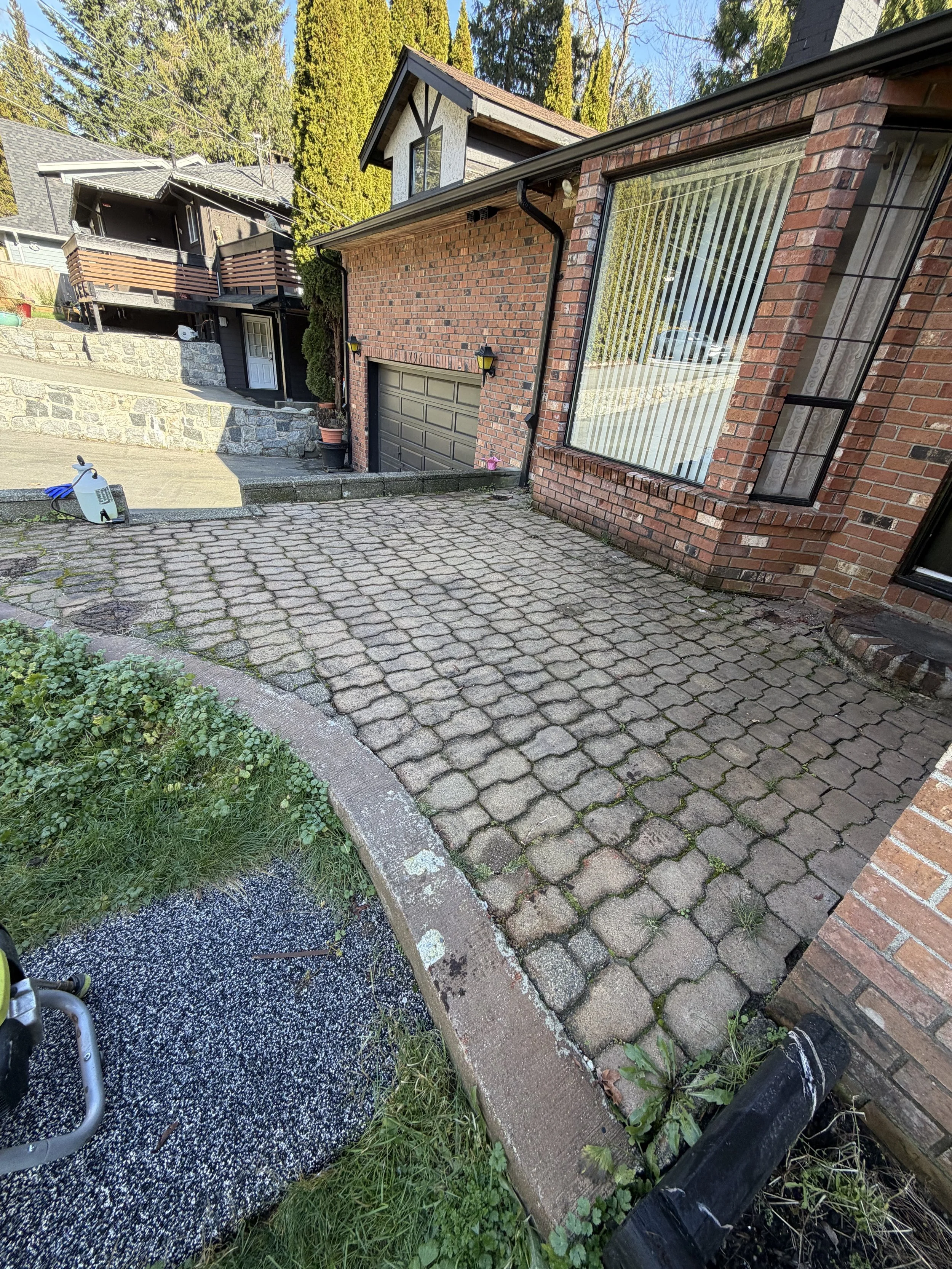View of a brick house with a large window, a garage door, and a small driveway made of interlocked bricks. The driveway slopes downward, with a curved garden bed with some grass and weeds on the left. There is a black drainage pipe on the right side of the image and a small piece of outdoor equipment on the left.