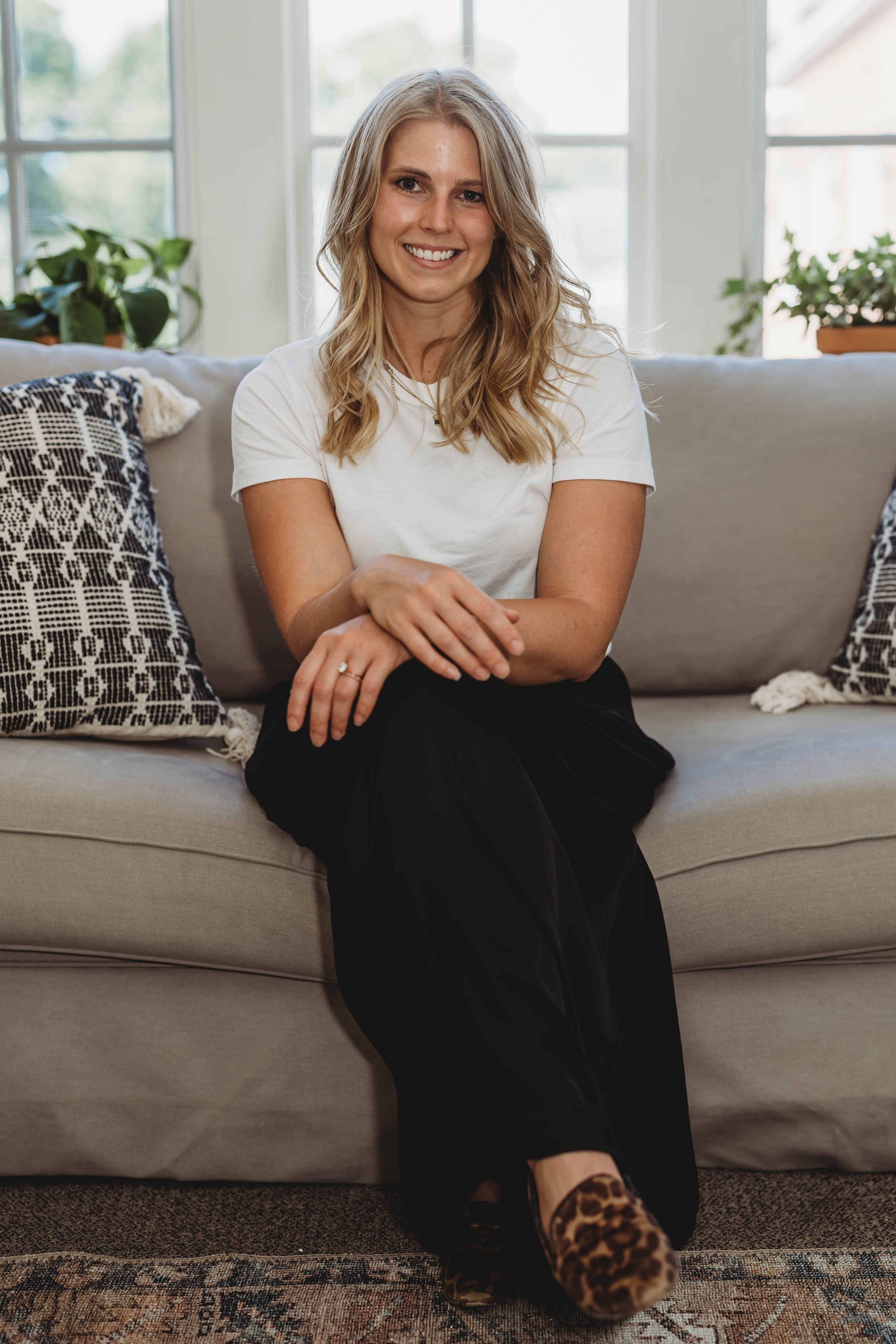 A woman sitting on a beige couch, smiling, with blonde wavy hair, wearing a white t-shirt and black pants. She has leopard print shoes and colorful patterned pillows nearby. The background includes large windows with greenery outside.