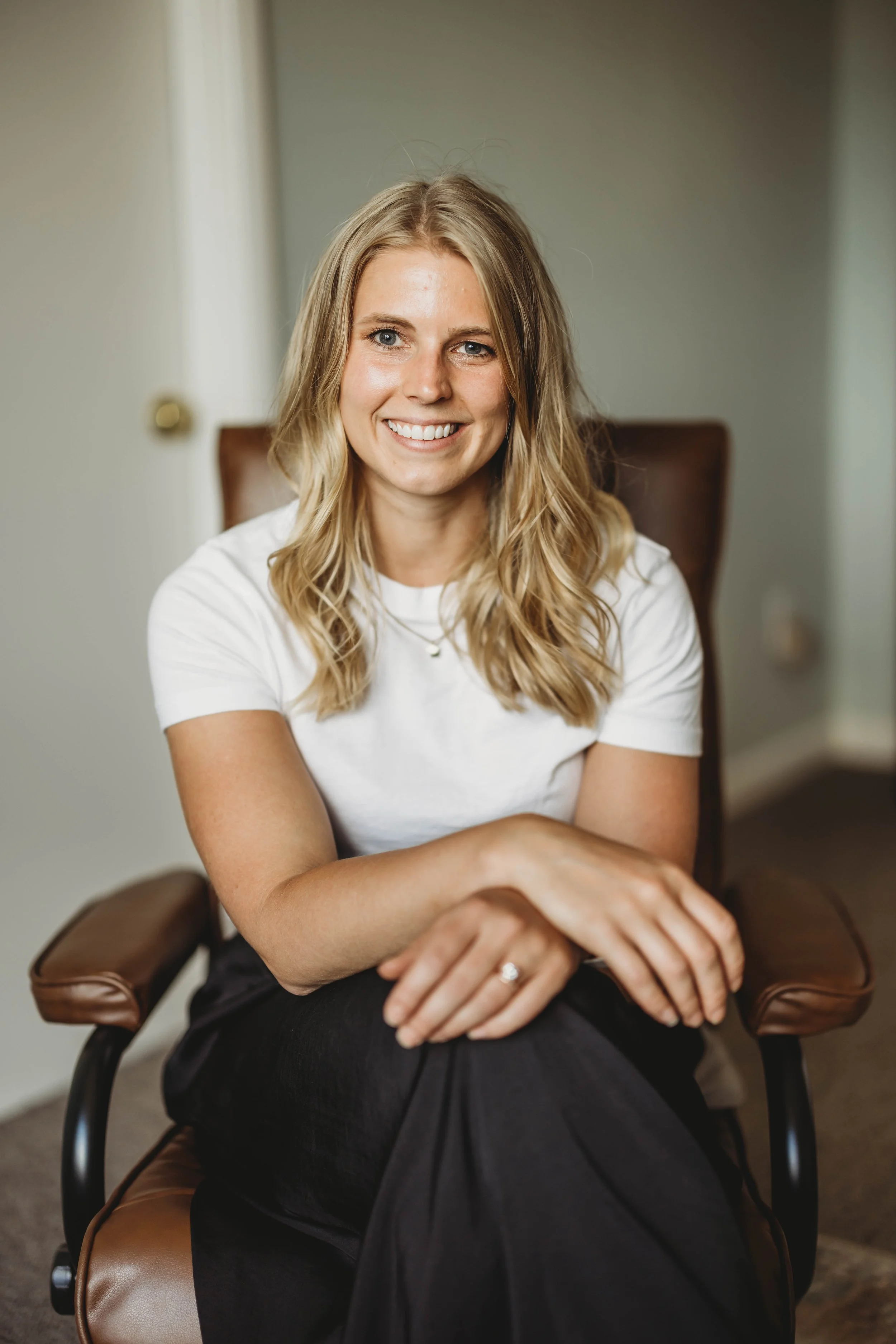 A young woman with blonde, wavy hair smiling and sitting in a brown leather chair with armrests. She is wearing a white t-shirt and black pants, with her arms crossed on her lap.