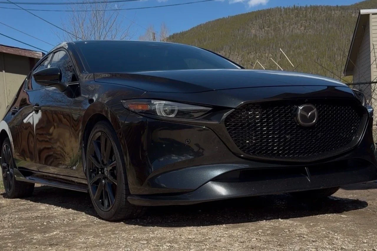 A black Mazda sedan parked outdoors on a dirt surface, with a house and a hill with trees in the background under a clear blue sky.
