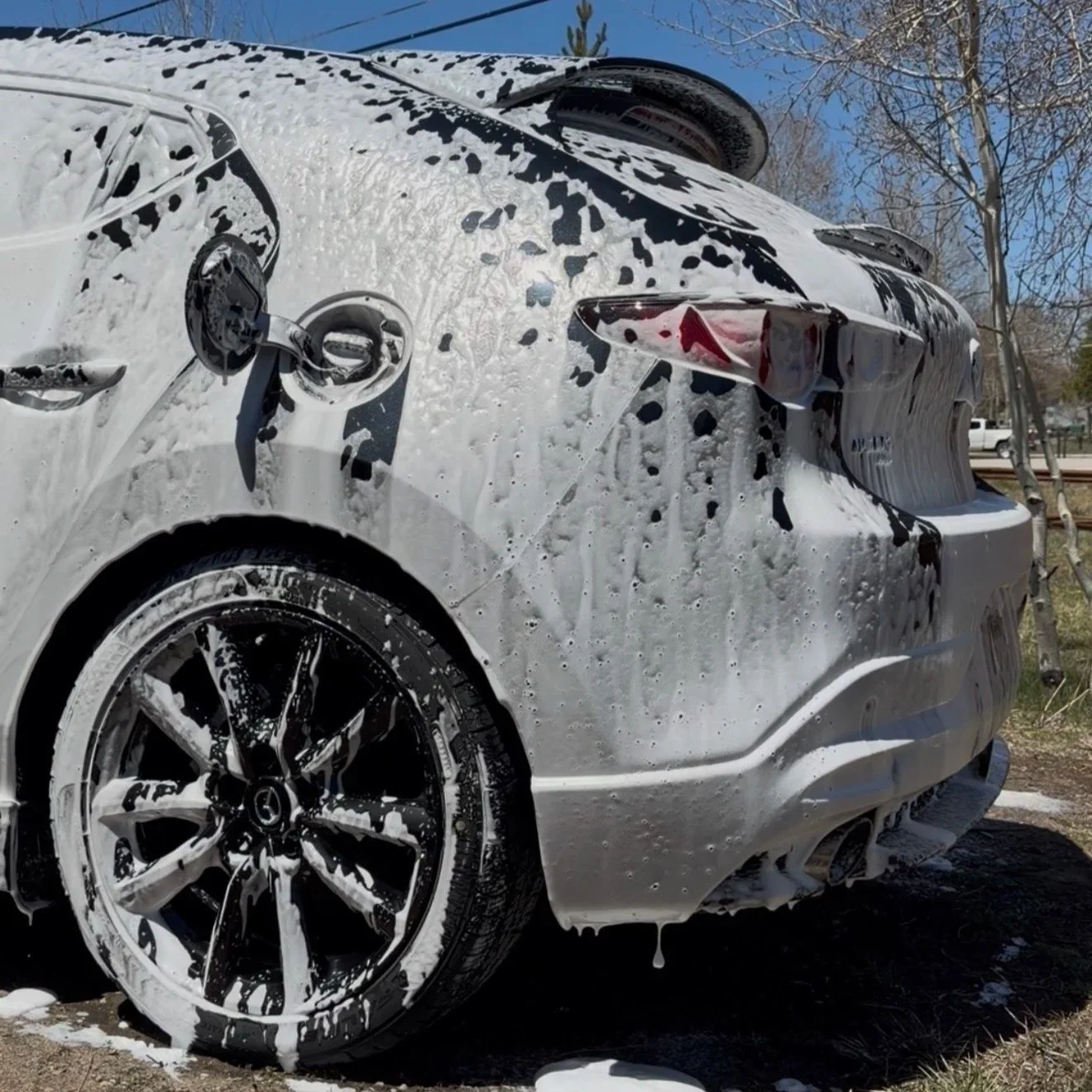 White sedan car covered in soap suds after washing, with foam on the rear and side, parked outdoors with trees in the background.