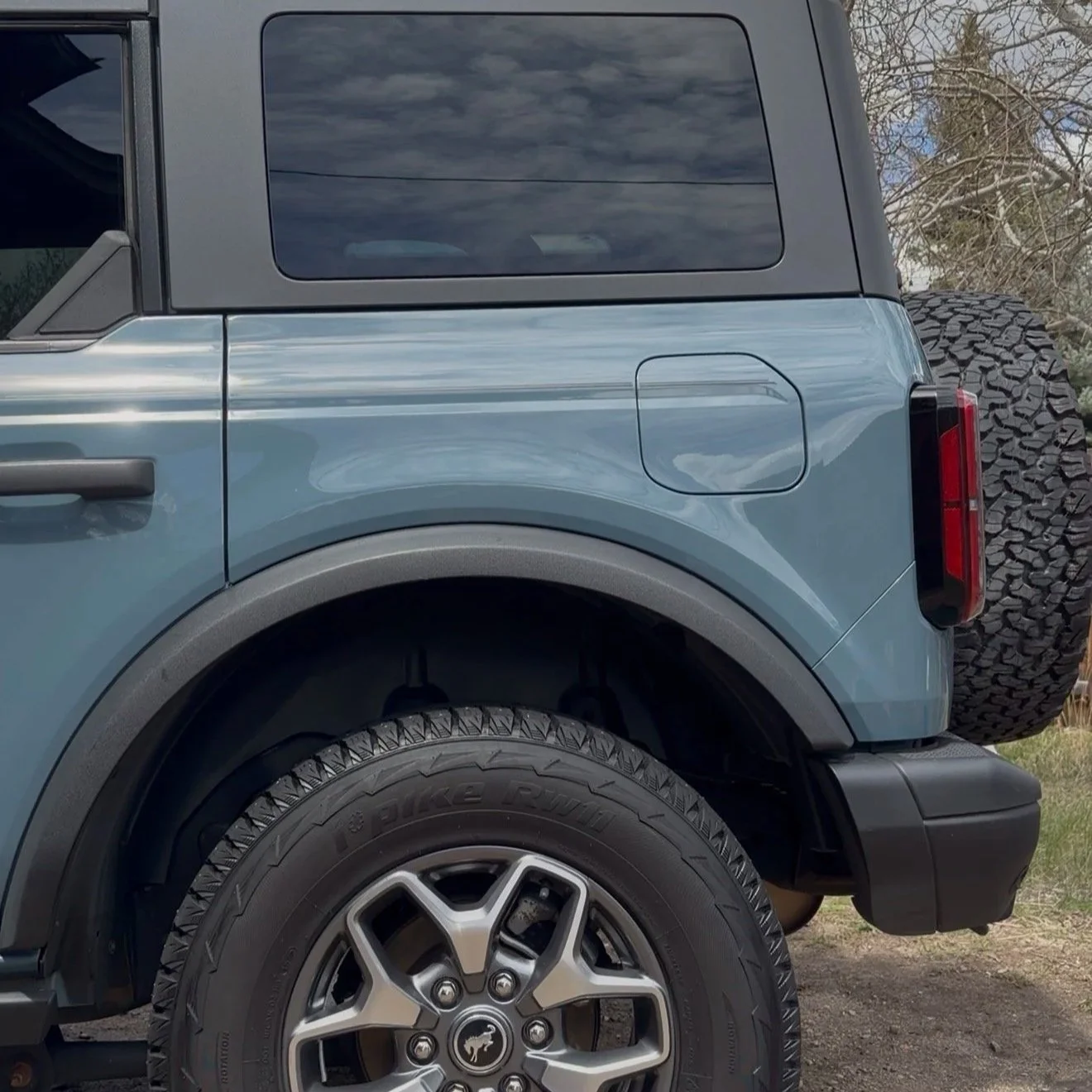 Close-up of the rear side of a blue off-road vehicle with large tires, a spare tire at the back, and tinted windows.