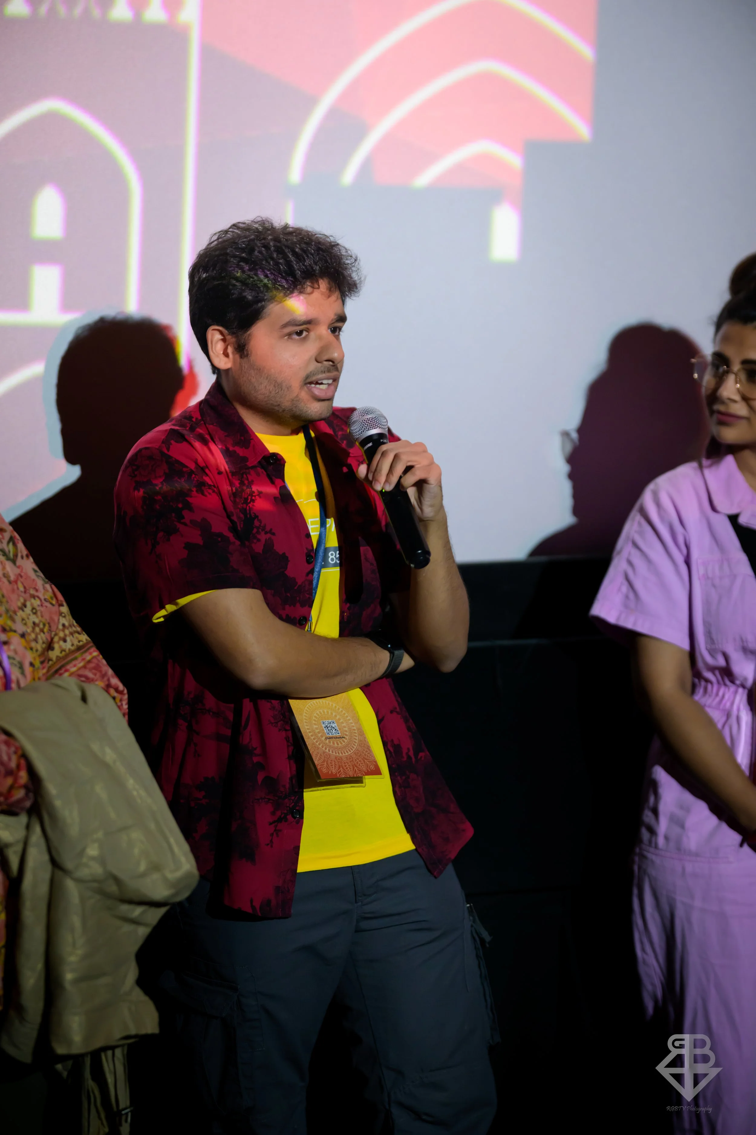 A young man holding a microphone and speaking at an event, with people and a projected screen in the background.