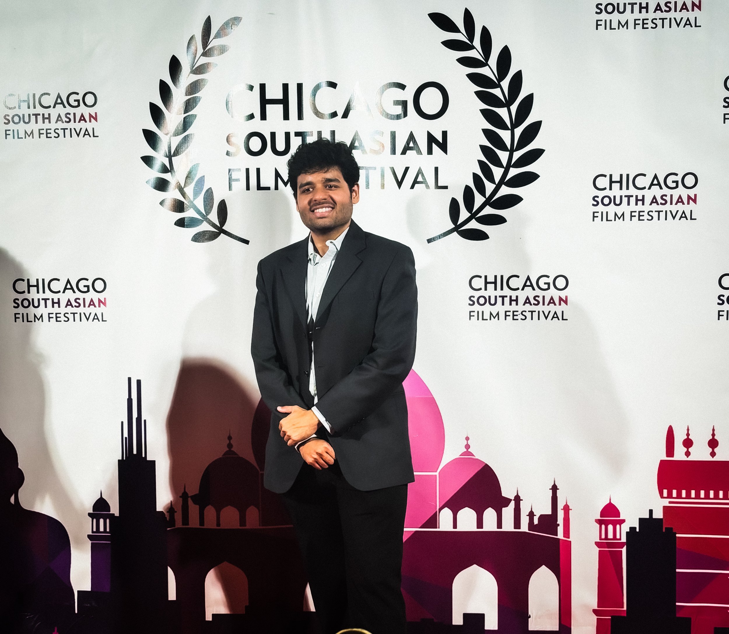 A young man in a black suit and white shirt standing with his hands clasped in front of a backdrop at the Chicago South Asian Film Festival. The backdrop features the festival's logo, Arabic-inspired architectural silhouettes, and the event's name.