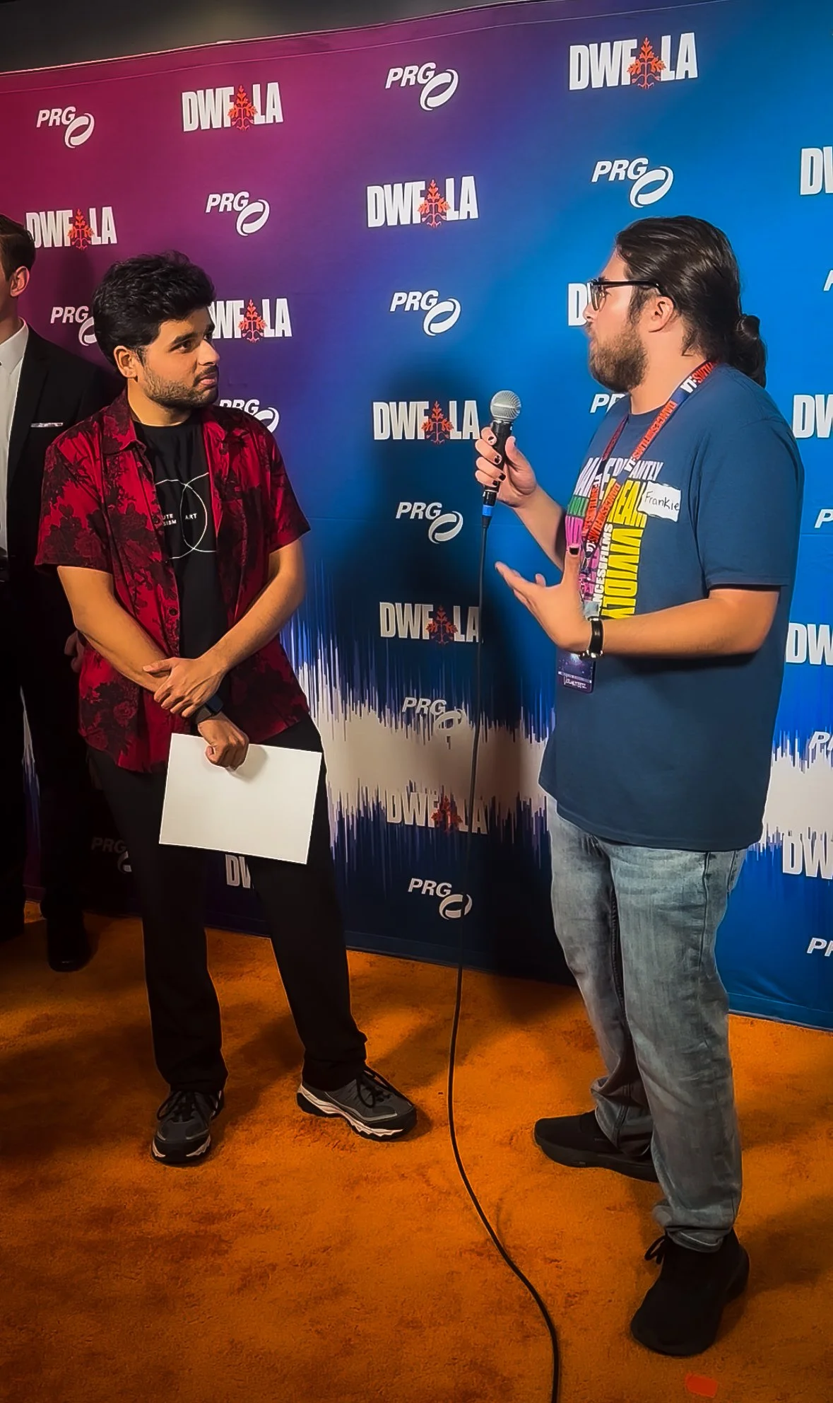 Two men having a conversation at an event, with a blue and purple backdrop featuring logos and the text "DWF LA" and "PRG." One man wears a patterned red and black shirt and holds a piece of paper, while the other man, wearing glasses and a blue T-shirt with colorful text, speaks into a microphone.