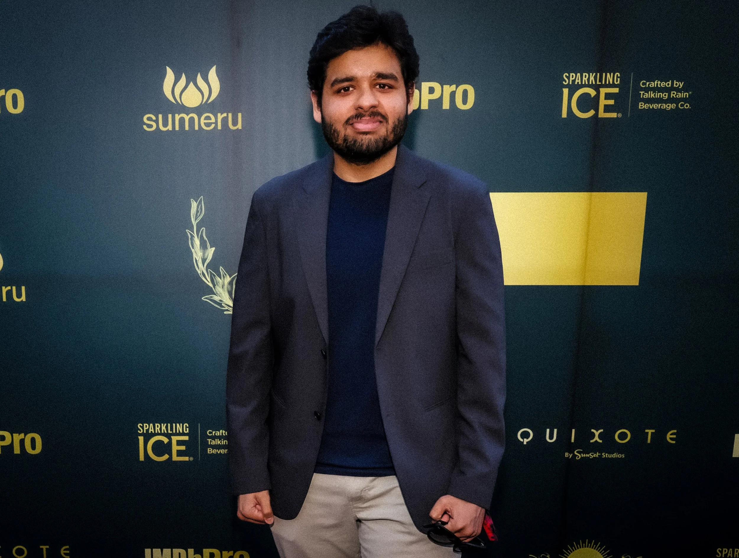 A man in a dark blazer and beige pants standing in front of a dark green backdrop with gold logos and text, smiling at the camera.