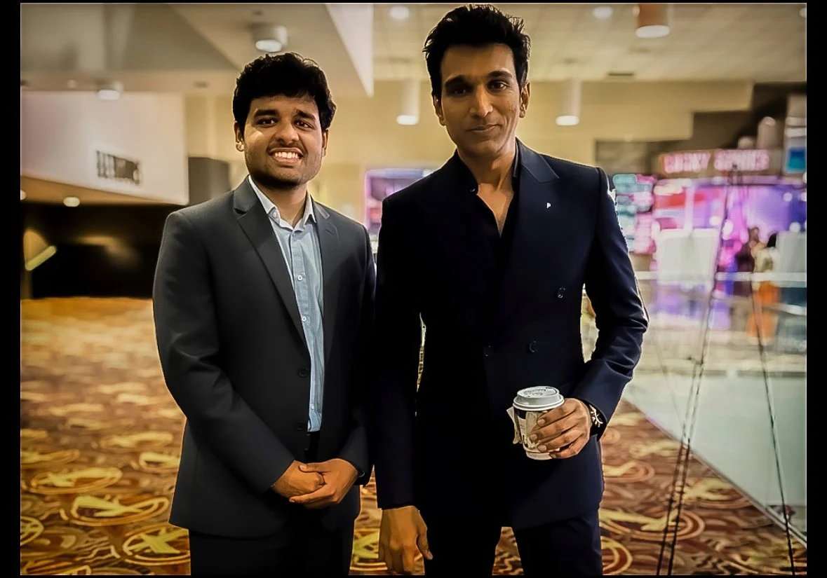 Two men in formal suits standing inside a shopping mall or conference venue, one holding a coffee cup, posing for the camera with a blurred background.