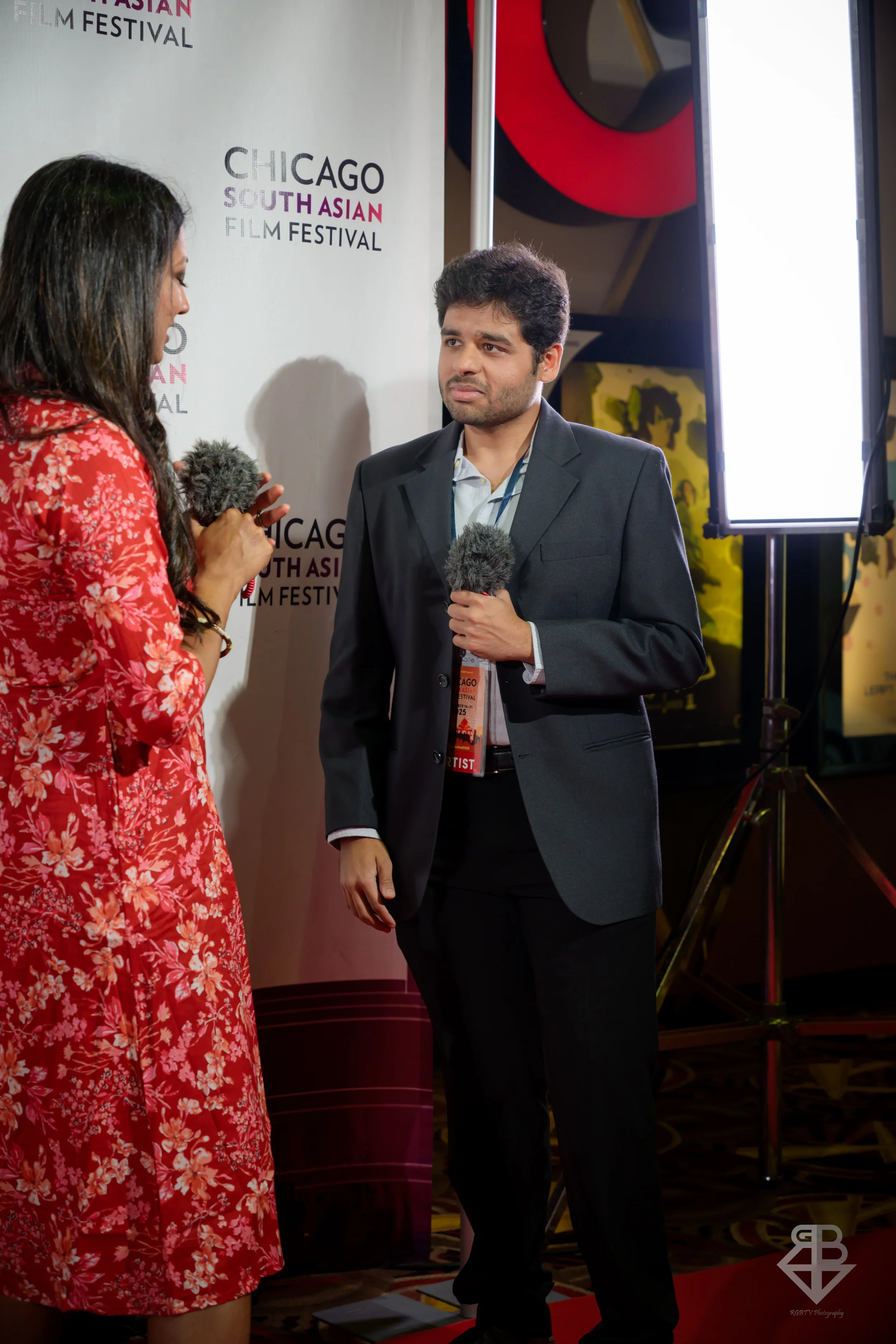 A man being interviewed at the Chicago South Asian Film Festival, standing near a woman holding a microphone, with a backdrop displaying the festival's logo.