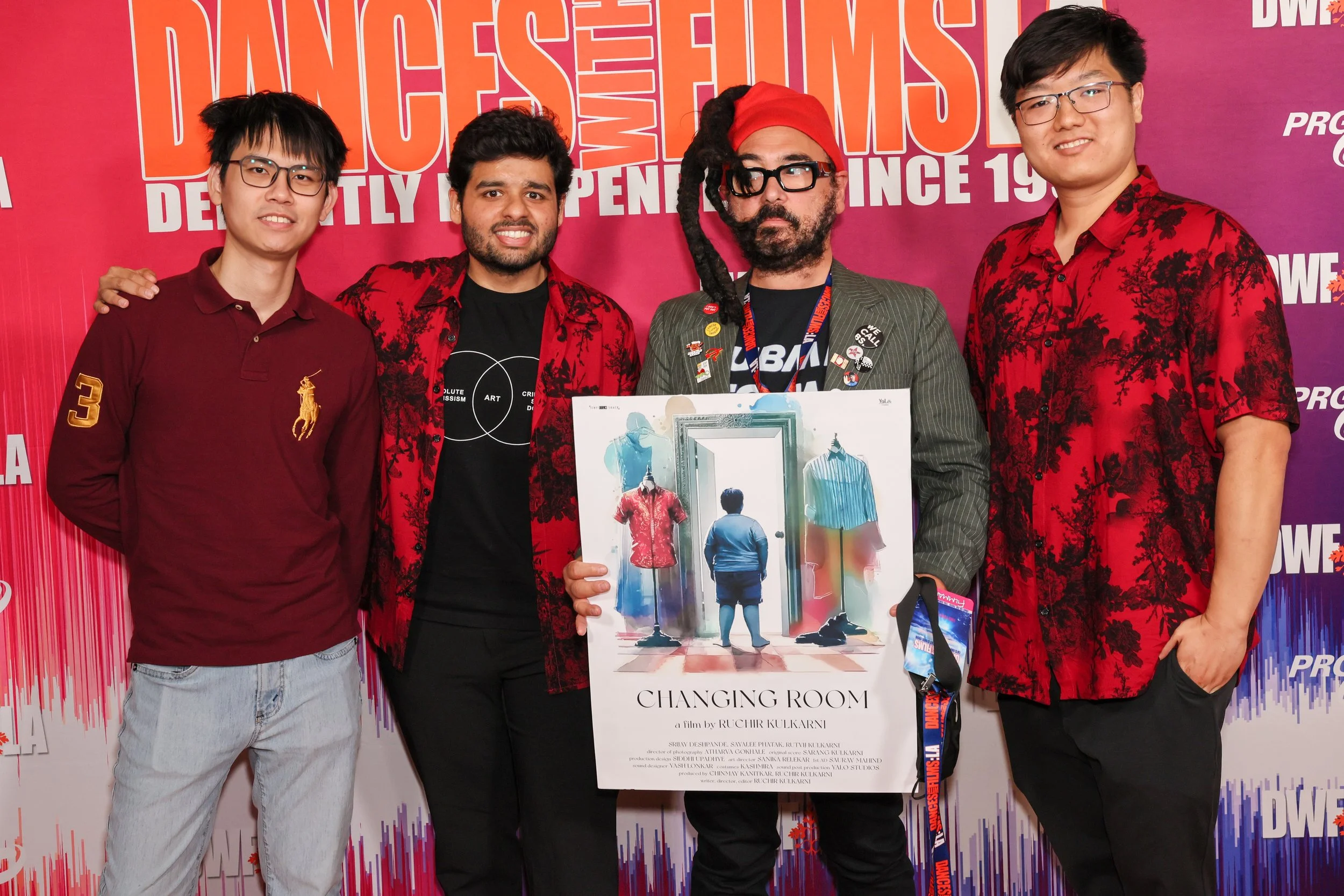 Five men pose together at a film event, with three wearing red and black outfits, one holding a poster for the film 'Changing Room', and a pink backdrop with text related to the event.
